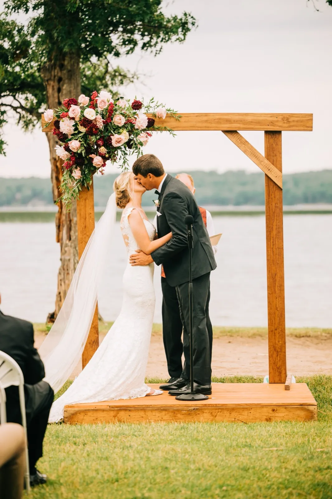 Shannon and Zach share their first kiss beneath a wooden arch trimmed with pink and burgundy florals, Gull Lake behind them at Madden's — Tim Larsen Photography, Brainerd Lakes MN
