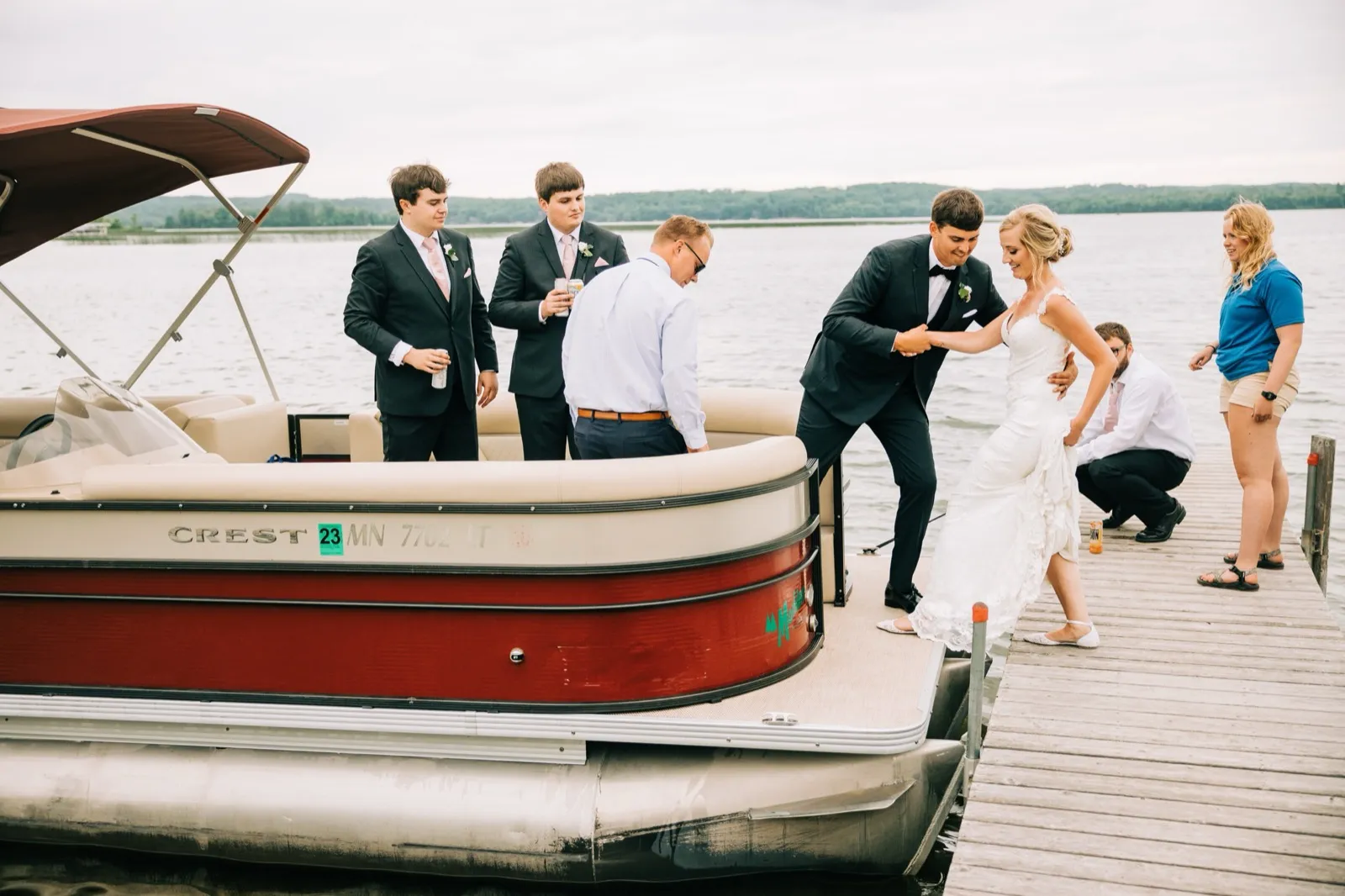 Zach steadies Shannon's hand as she steps from a wooden dock onto a red pontoon boat on Gull Lake, the bridal party pitching in — Tim Larsen Photography, Brainerd Lakes MN