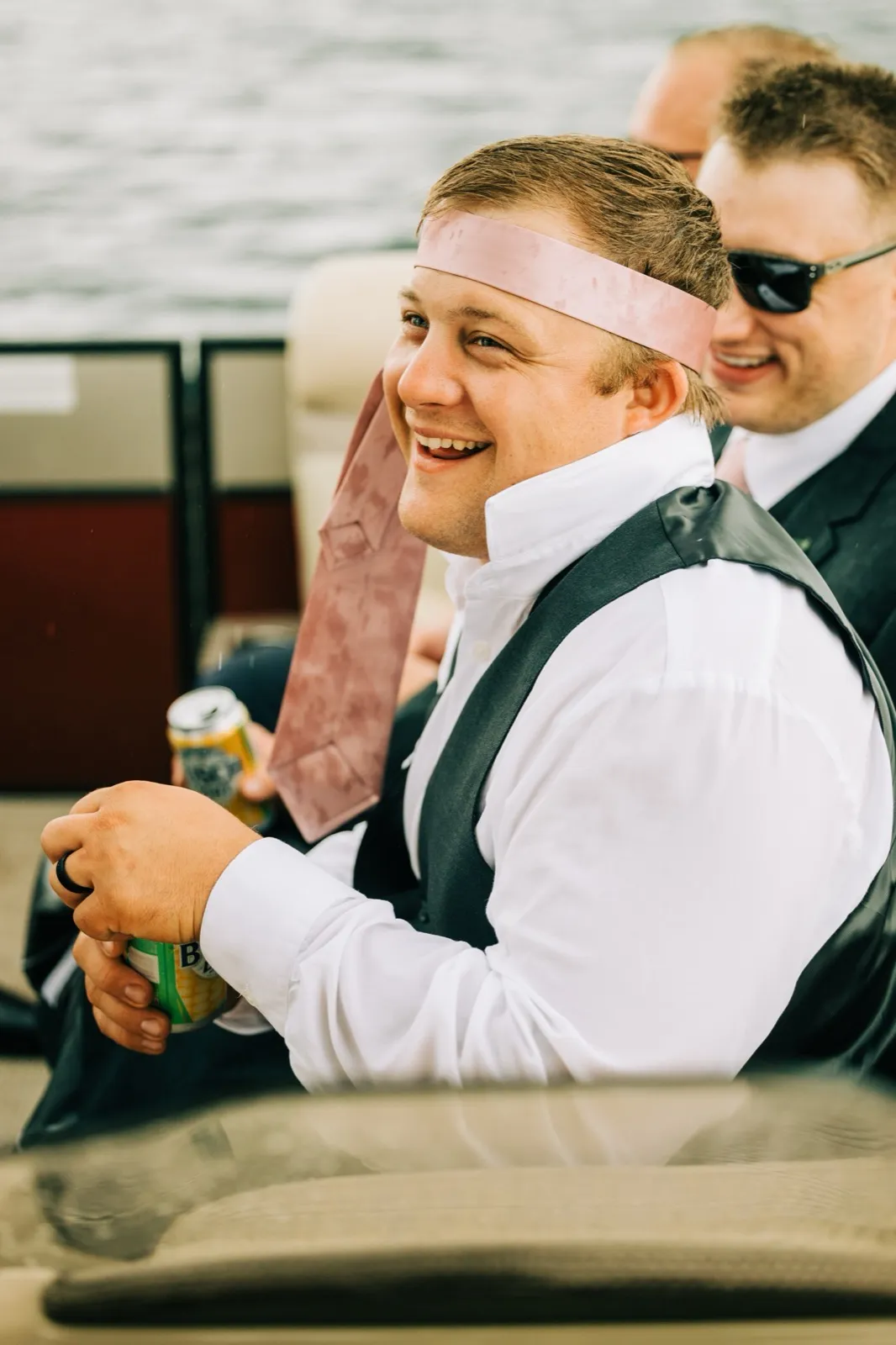 Zach laughs on the pontoon with his pink tie knotted around his head like a headband, a groomsman beside him — Tim Larsen Photography, Brainerd Lakes MN