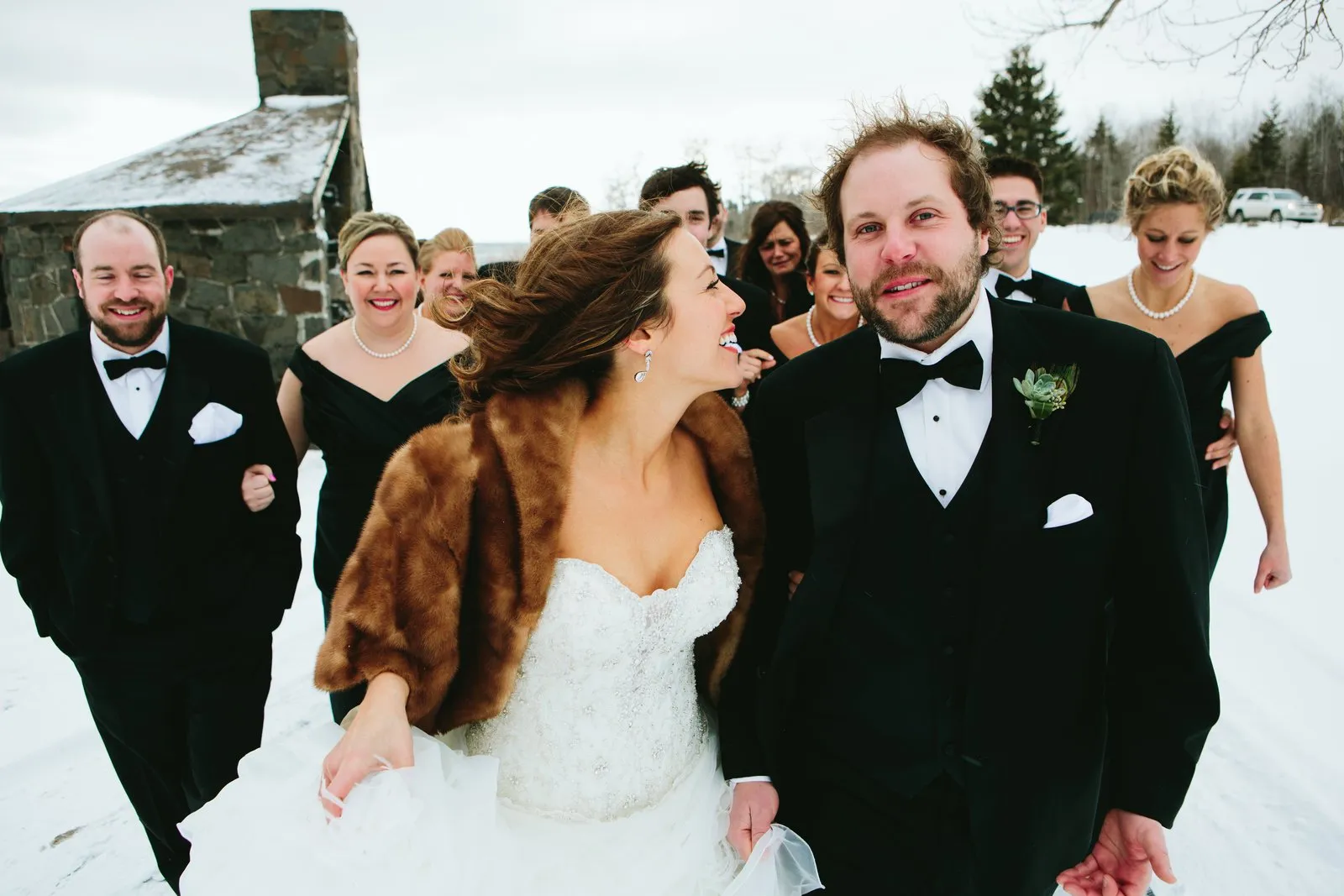 Bridal party walking toward camera with the bride laughing at the groom mid-stride beside a snowy stone ruin — Duluth winter wedding — Tim Larsen Photography, Duluth MN