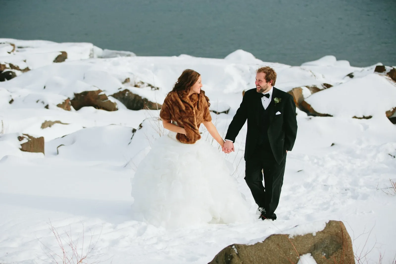 Bride in a fur stole leading the groom by the hand across the snow-covered Lake Superior shoreline — winter portrait in Duluth — Tim Larsen Photography, Duluth MN