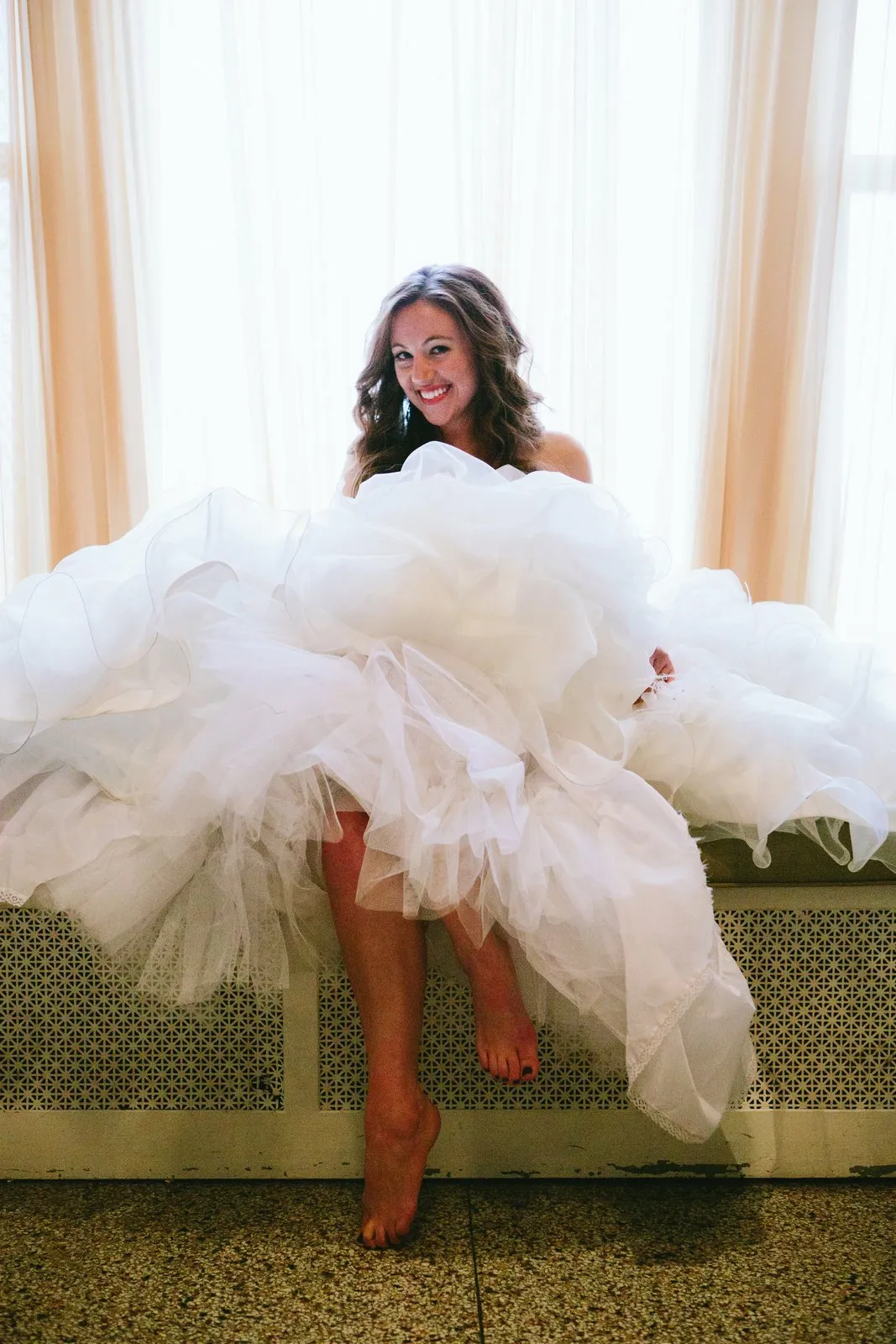 Bride seated on the radiator bench at a window, gown cascading and bare feet dangling, laughing — Greysolon Plaza bridal suite — Tim Larsen Photography, Duluth MN