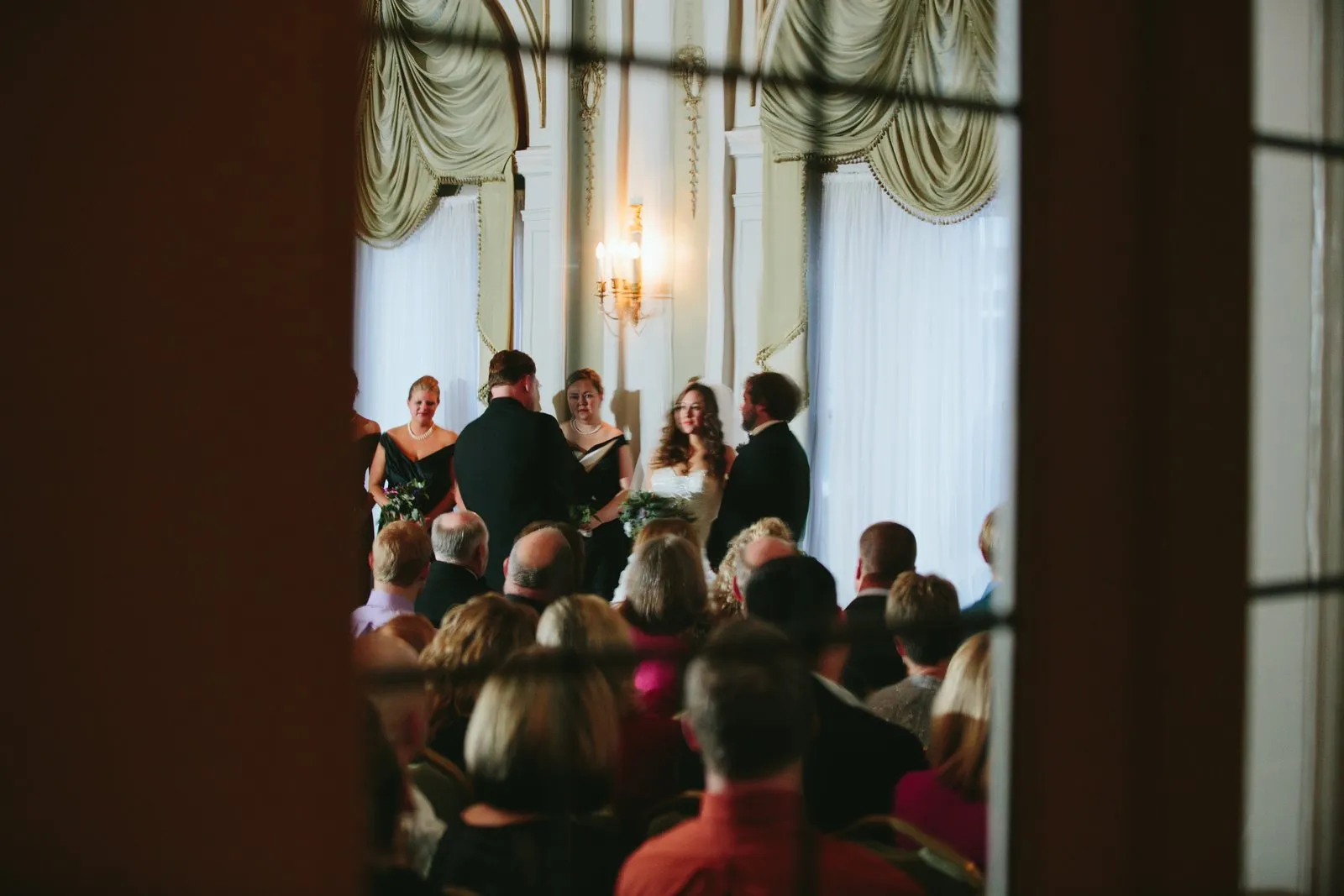 Ceremony framed through a doorway by dark architectural negative space, guests in the foreground — Greysolon Ballroom altar — Tim Larsen Photography, Duluth MN