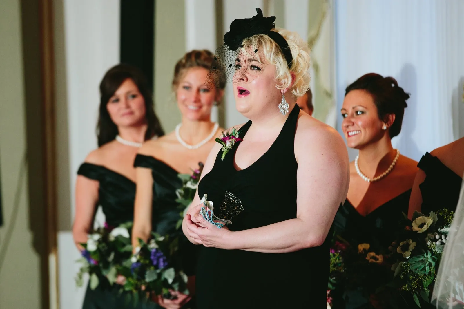 Bridesmaid in a black gown and fascinator mid-emotional-reaction during the ceremony, bridesmaids flanking in soft focus — Greysolon Ballroom — Tim Larsen Photography, Duluth MN