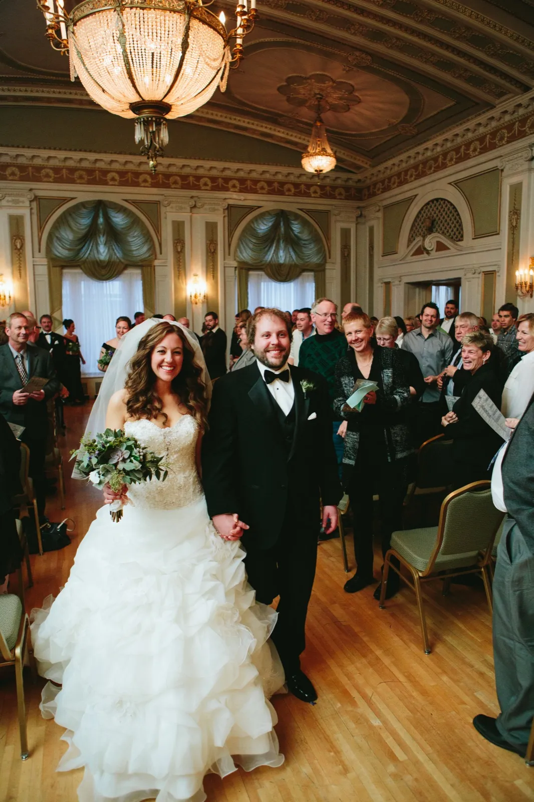 Joyful recessional down the Greysolon Ballroom aisle under the grand chandelier — bride and groom hand in hand, guests applauding — Tim Larsen Photography, Duluth MN