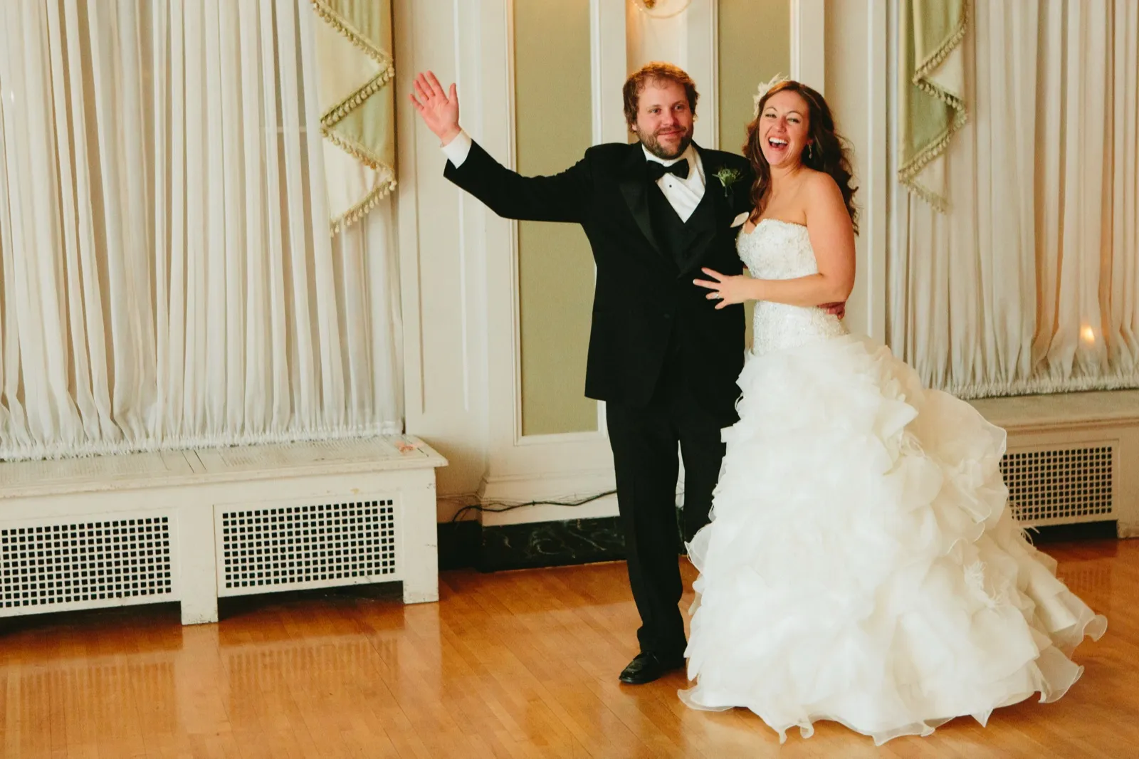 Grand entrance — the couple framed between draped windows on the ballroom dance floor, arms thrown wide laughing — Tim Larsen Photography, Duluth MN