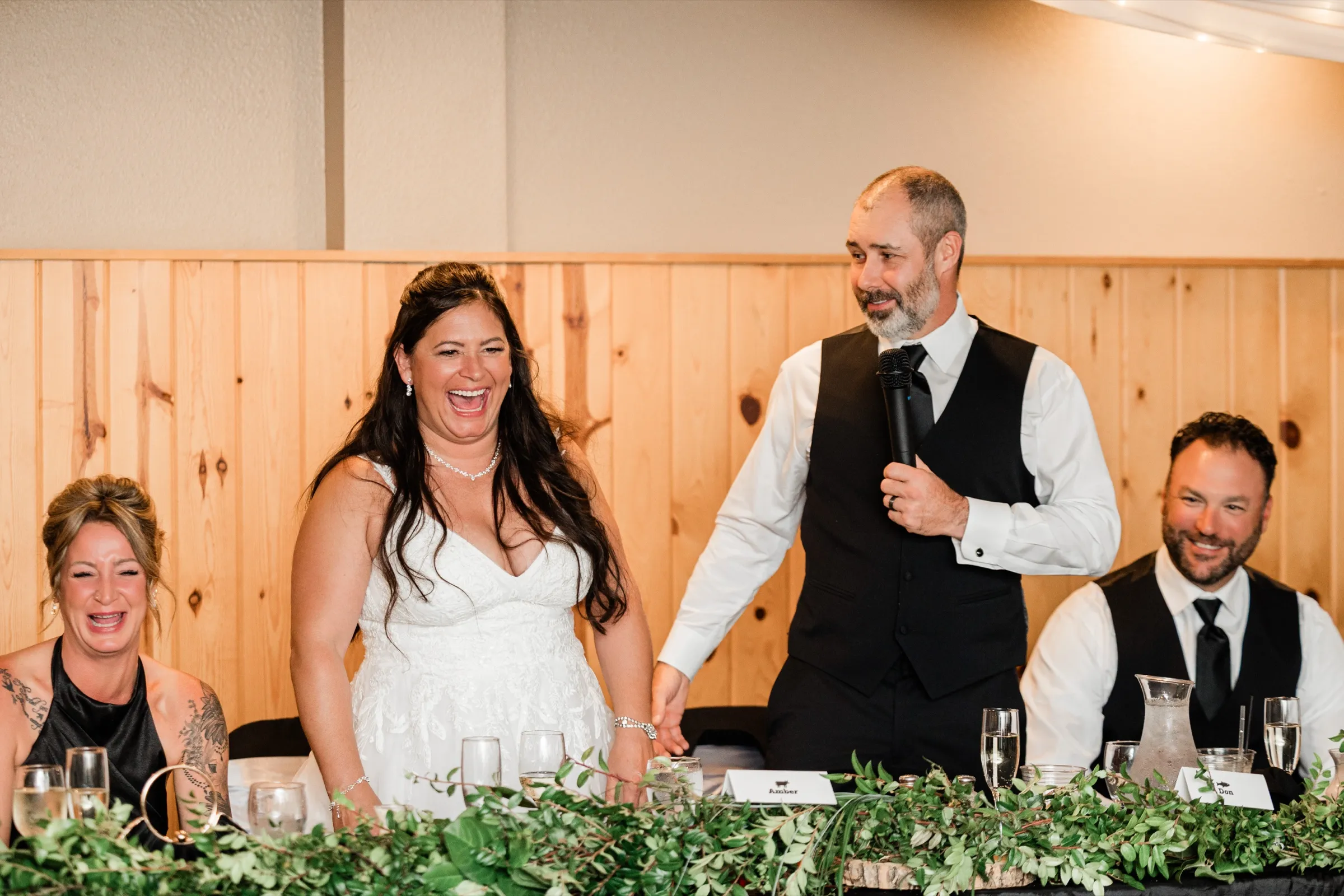Amber laughs with her mouth wide open as Don speaks into a microphone, while a bridesmaid at the head table laughs with her eyes shut. — Tim Larsen Photography, Brainerd Lakes MN