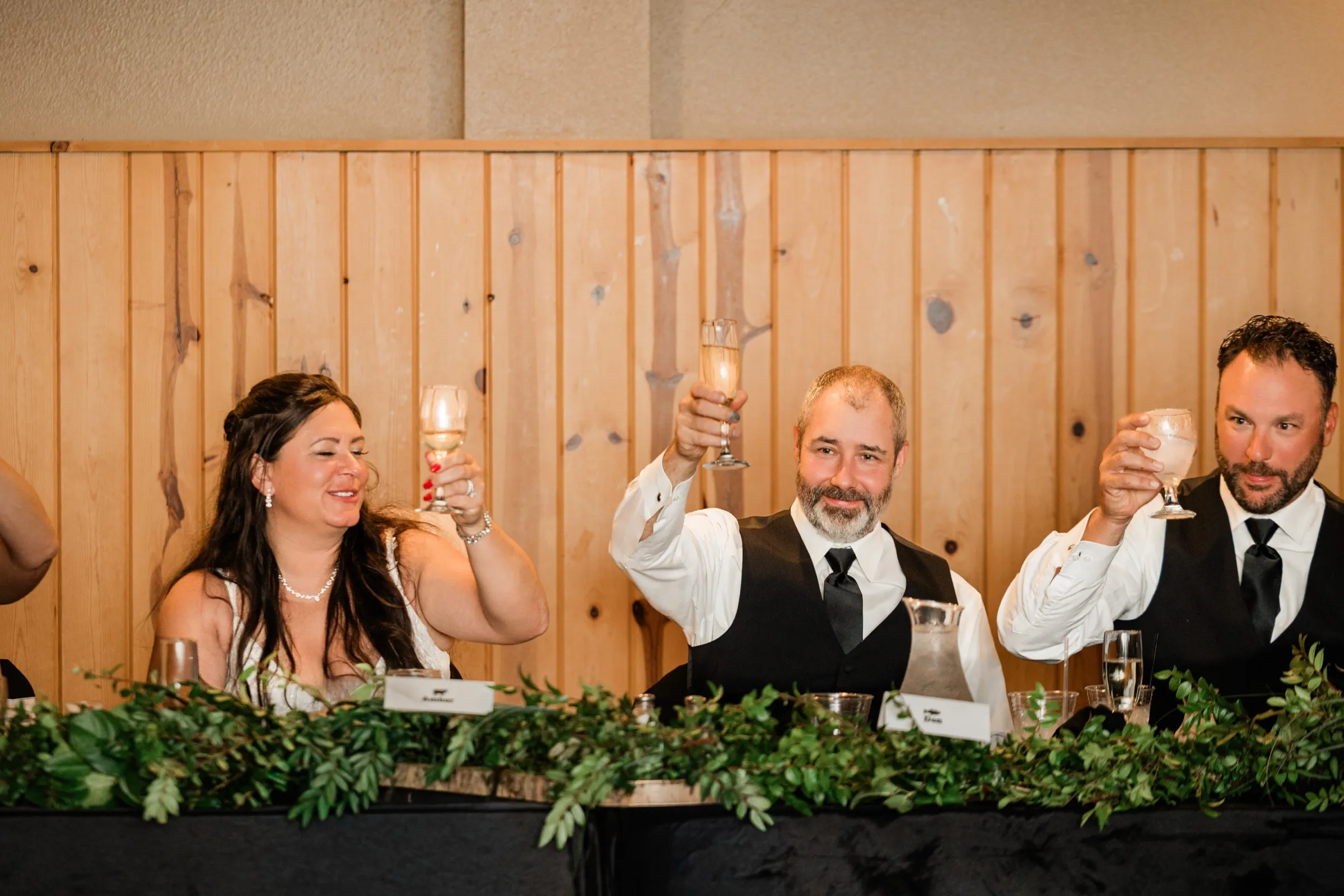 Amber, Don and a groomsman raise their glasses at a head table lined with a greenery garland in front of a knotty pine wall. — Tim Larsen Photography, Brainerd Lakes MN