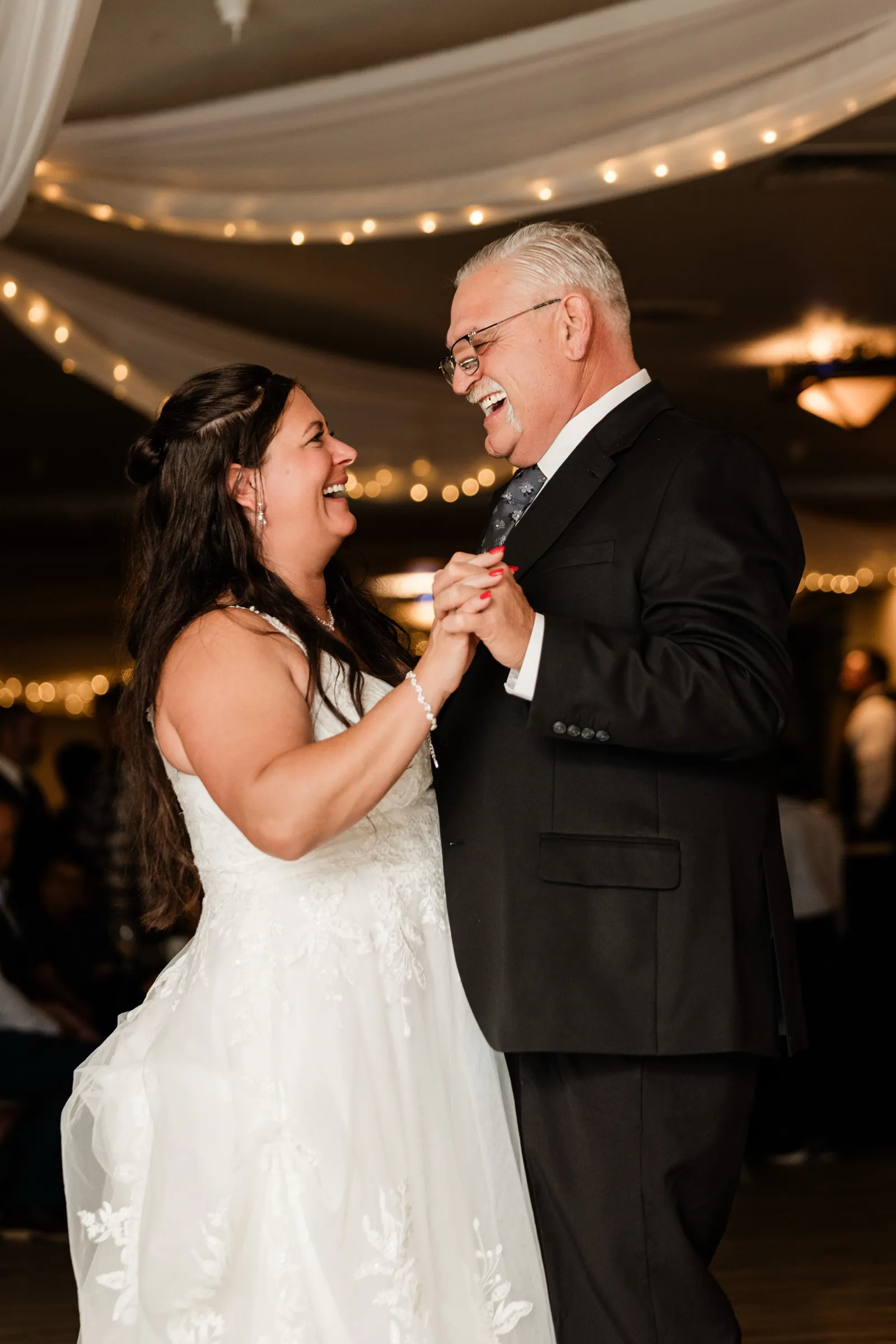 Amber and an older man in a dark suit and glasses laugh face to face as they dance holding hands under string lights. — Tim Larsen Photography, Brainerd Lakes MN