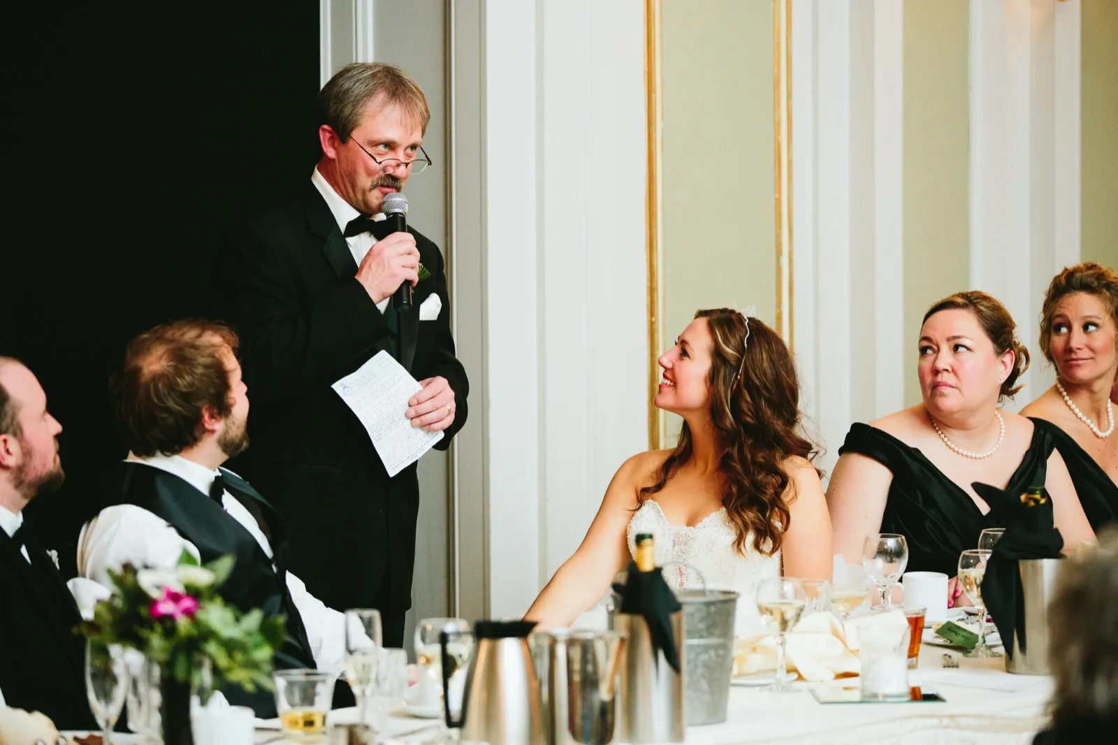 Father of the bride at the mic mid-toast, the bride looking up adoringly with guests' reactions flanking — Greysolon Ballroom speeches — Tim Larsen Photography, Duluth MN