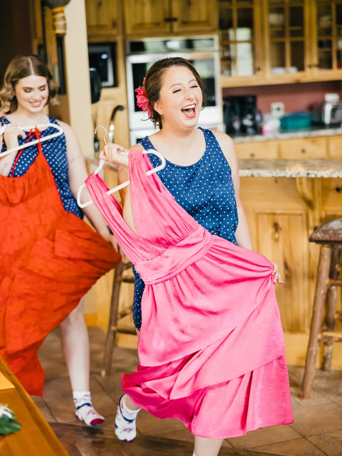 The bride laughs with her mouth wide open while holding a hot pink dress on a hanger, with a bridesmaid in a navy polka-dot dress holding a red dress beside her in a wood-paneled kitchen. — Tim Larsen Photography, Brainerd Lakes MN