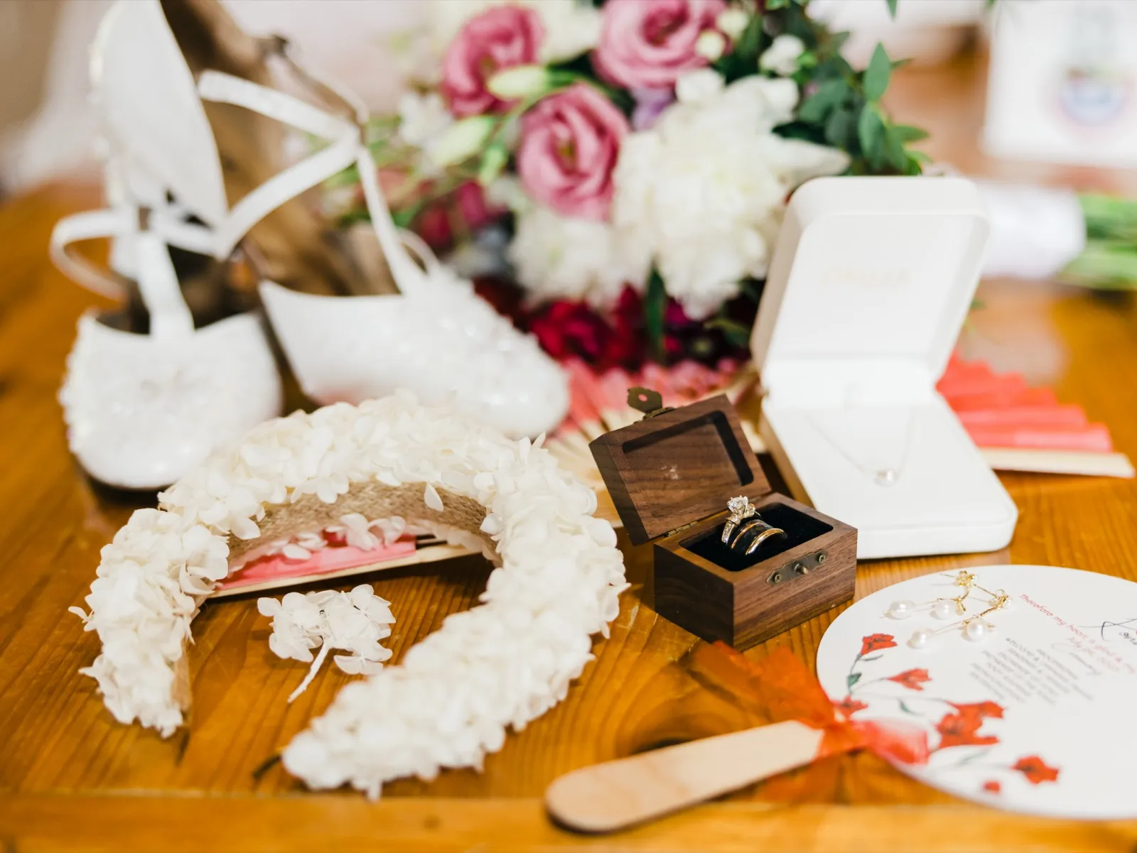 A flat-lay arrangement on a wood table showing white bridal heels, a floral hair piece, the bride's bouquet, an open ring box with stacked rings, a necklace in a jewelry case, pearl earrings, and a coral-ribboned program. — Tim Larsen Photography, Brainerd Lakes MN