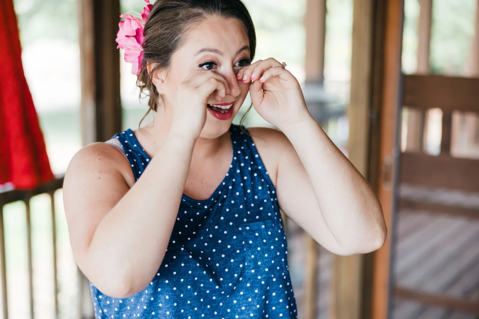 The bride laughs through tears as she wipes her eyes, wearing a polka-dot tank top and a pink flower in her hair on a covered porch. — Tim Larsen Photography, Brainerd Lakes MN