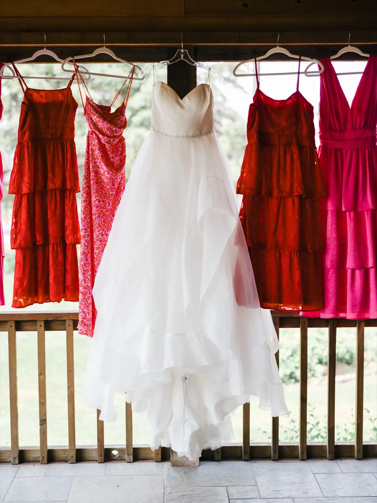 A white wedding gown hangs centered between four bridesmaid dresses in shades of red and pink along a wooden porch railing. — Tim Larsen Photography, Brainerd Lakes MN