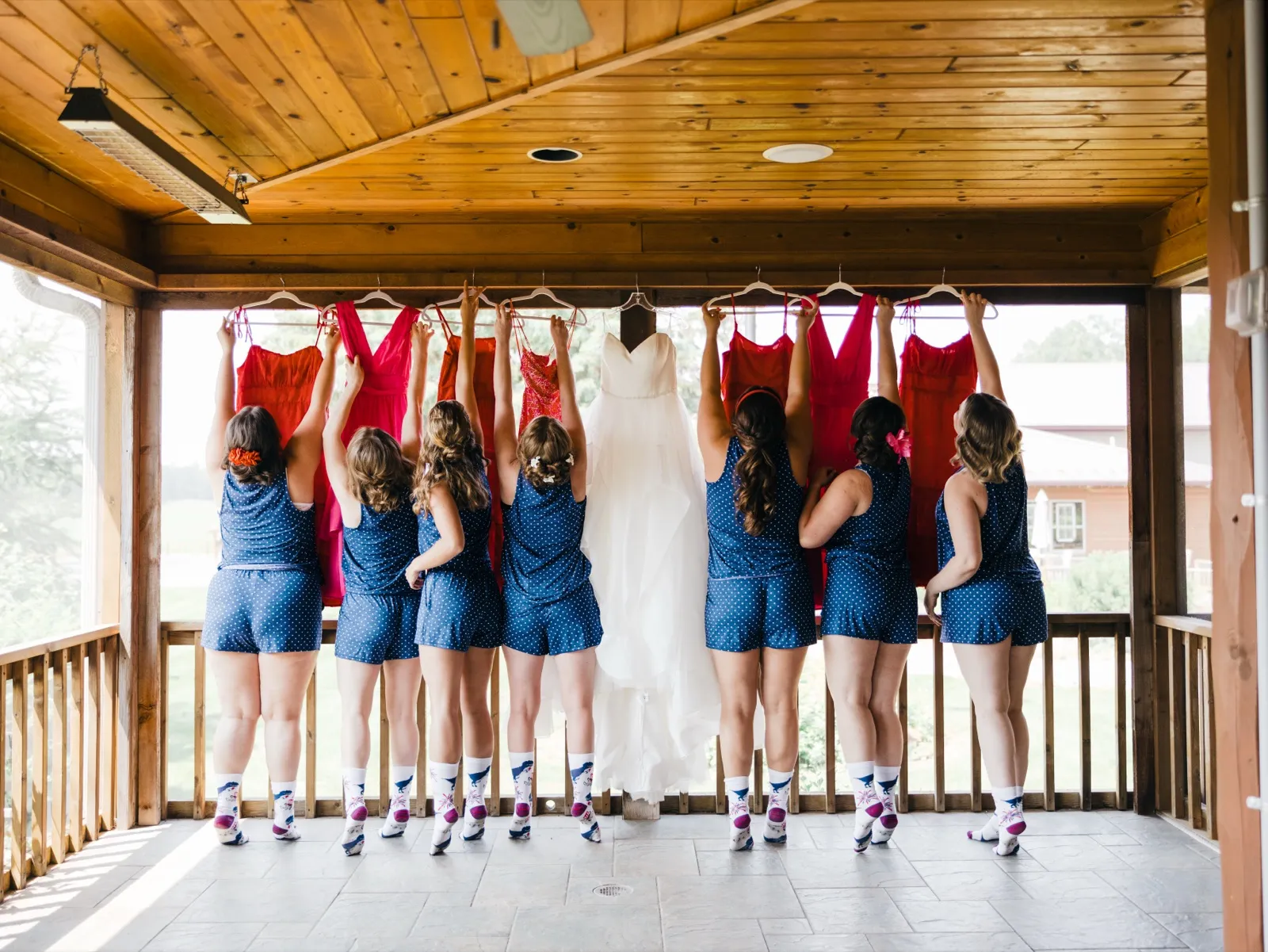 Seven bridesmaids in matching navy polka-dot rompers and patterned socks stand with their backs to camera on a covered wooden porch, holding red dresses and the bride's white gown overhead on hangers. — Tim Larsen Photography, Brainerd Lakes MN