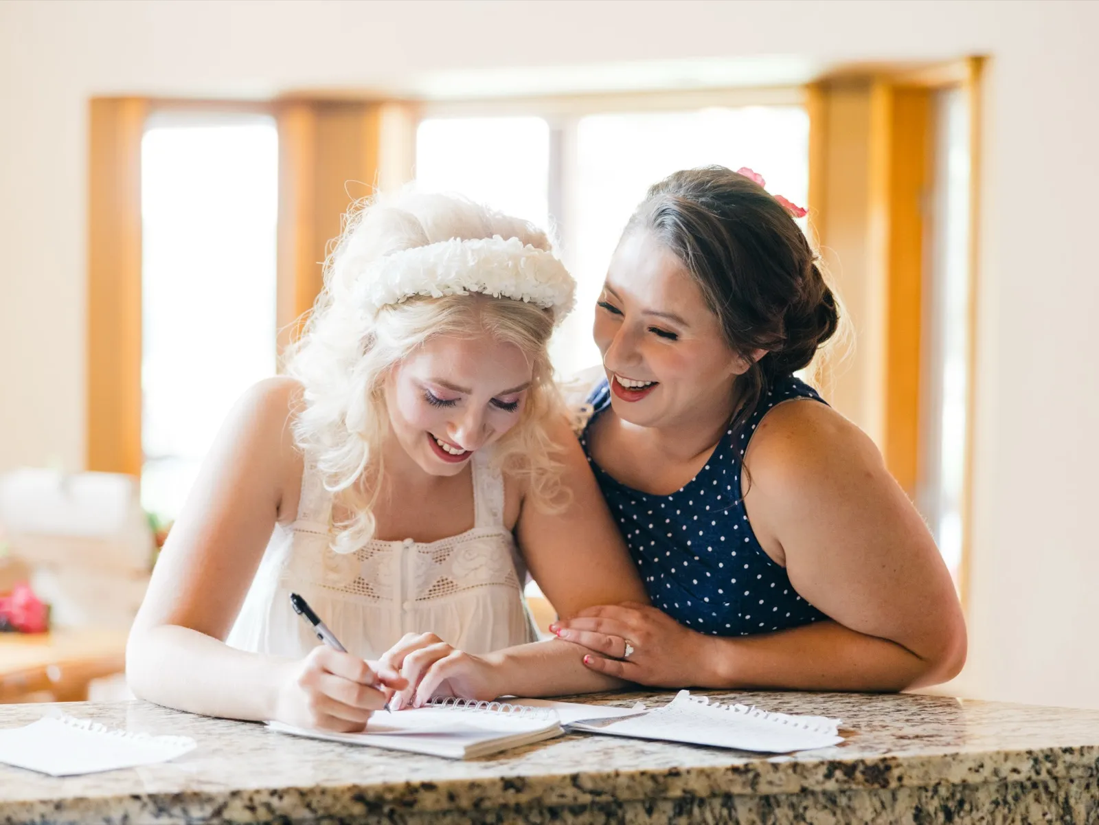 The bride, wearing a floral headpiece, writes in a notebook at a granite counter while a bridesmaid in a polka-dot dress leans in laughing beside her. — Tim Larsen Photography, Brainerd Lakes MN