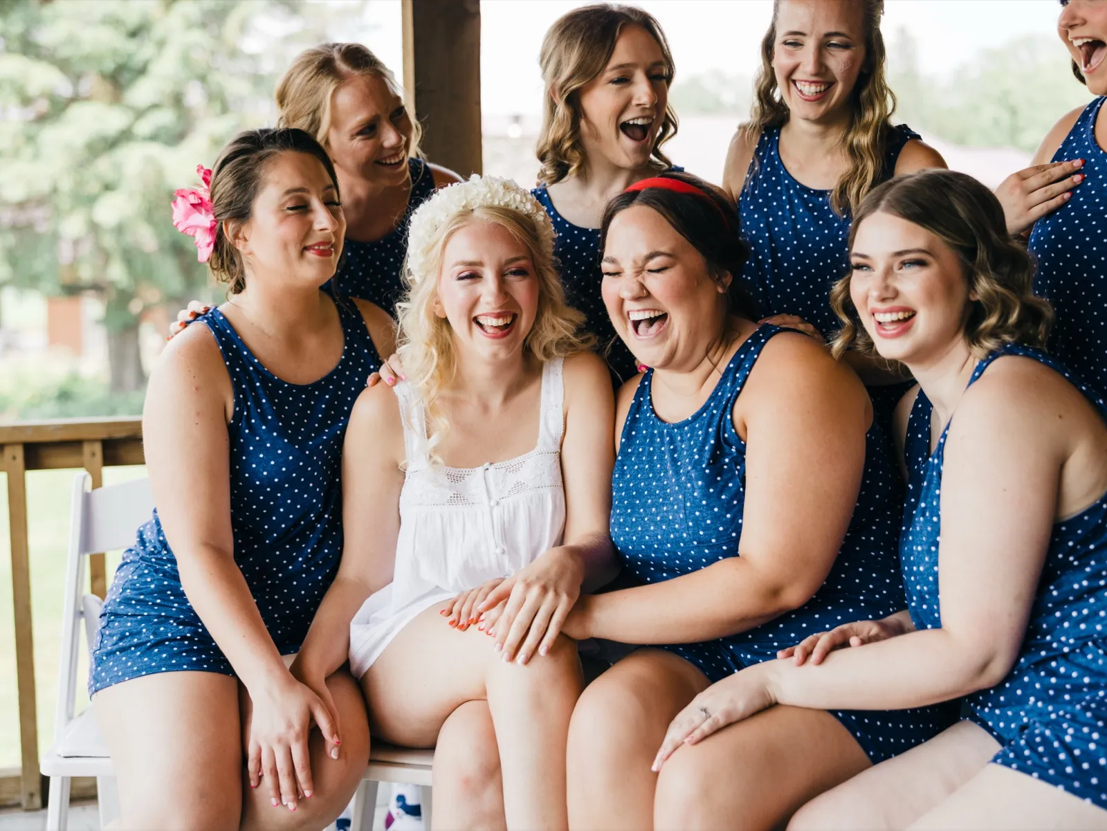 The bride in a white slip and floral crown laughs hard surrounded by bridesmaids in matching navy polka-dot rompers on a covered porch. — Tim Larsen Photography, Brainerd Lakes MN