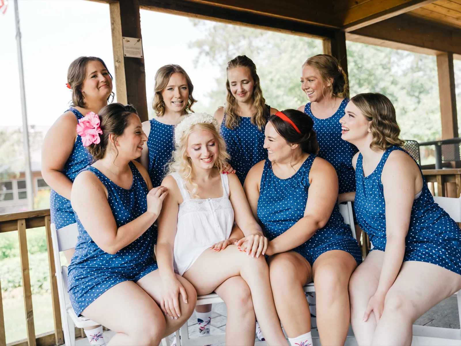 The bride sits on a bench in a white slip and floral crown surrounded by seven bridesmaids in matching blue polka-dot rompers, all laughing together on a covered porch. — Tim Larsen Photography, Brainerd Lakes MN