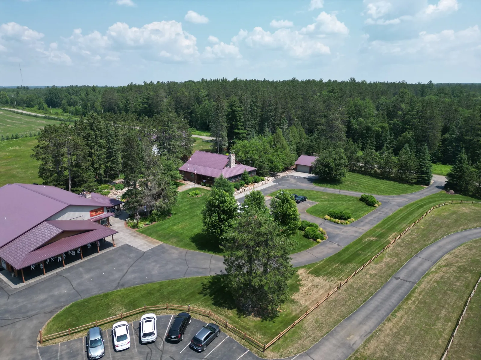 Aerial view of Pine Peaks Event Center showing the main lodge with red roof, an adjacent event barn, parked cars, and surrounding pine forest under a partly cloudy sky. — Tim Larsen Photography, Brainerd Lakes MN