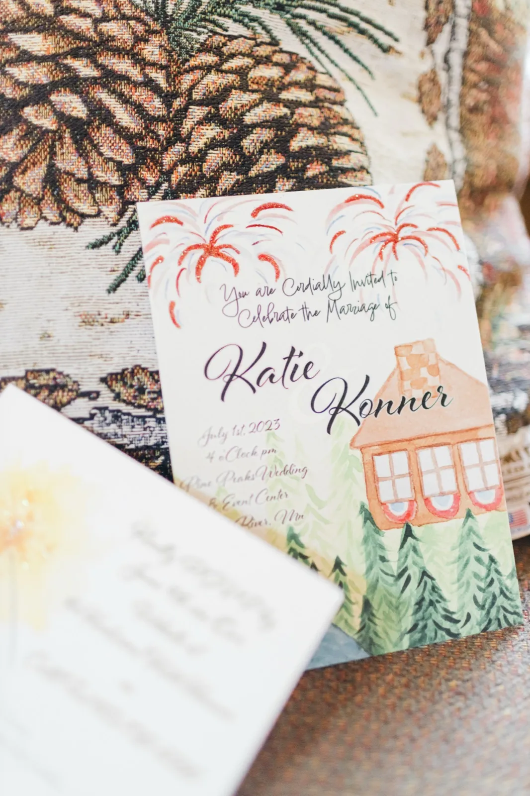 A hand-painted wedding invitation for Katie and Konnor featuring fireworks and a small cabin rests on a pinecone tapestry, with a second card visible in the foreground. — Tim Larsen Photography, Brainerd Lakes MN