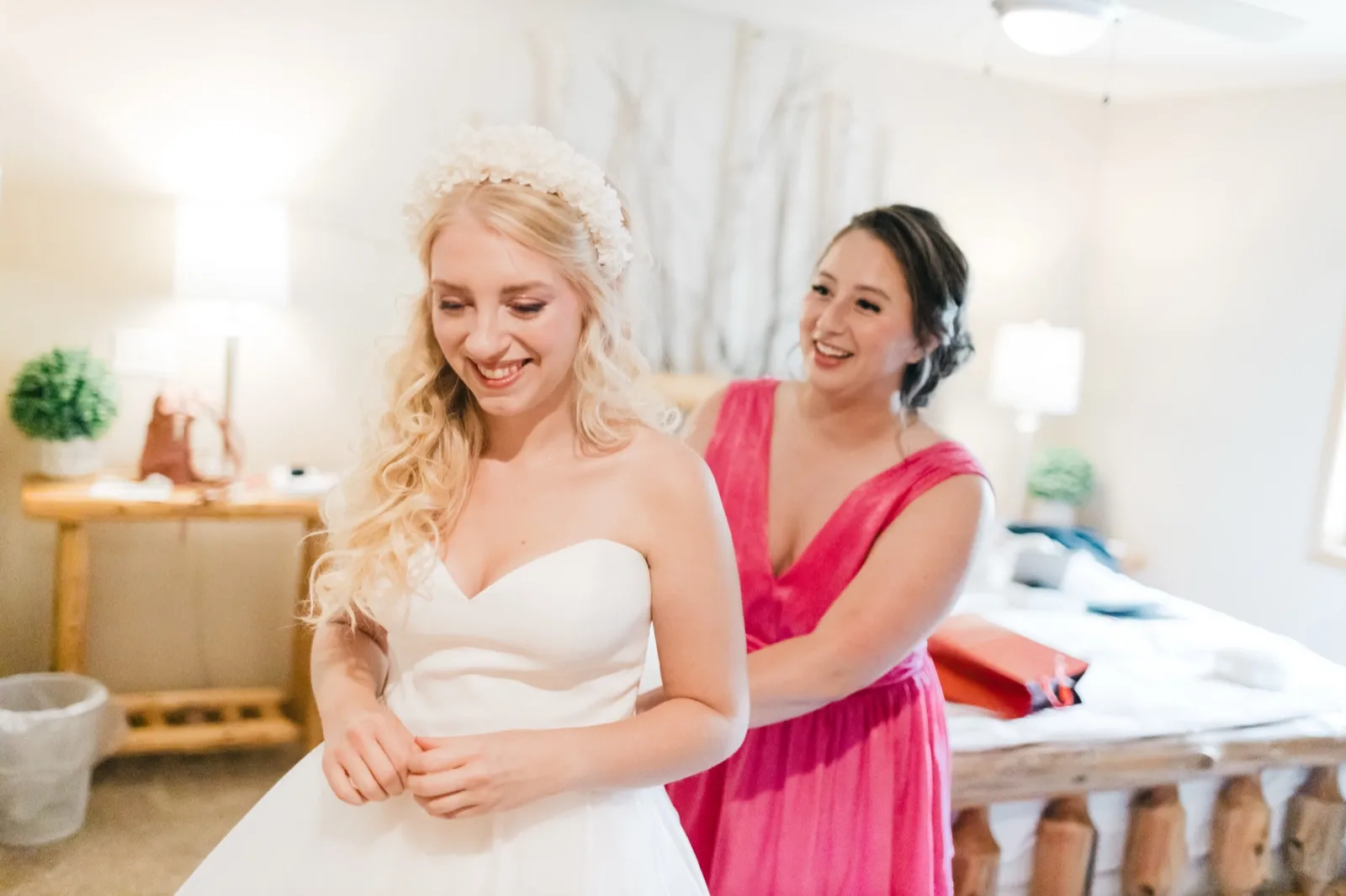 The bride laughs with her head tilted down while a bridesmaid in a pink dress fastens the back of her gown in a sunlit cabin bedroom. — Tim Larsen Photography, Brainerd Lakes MN