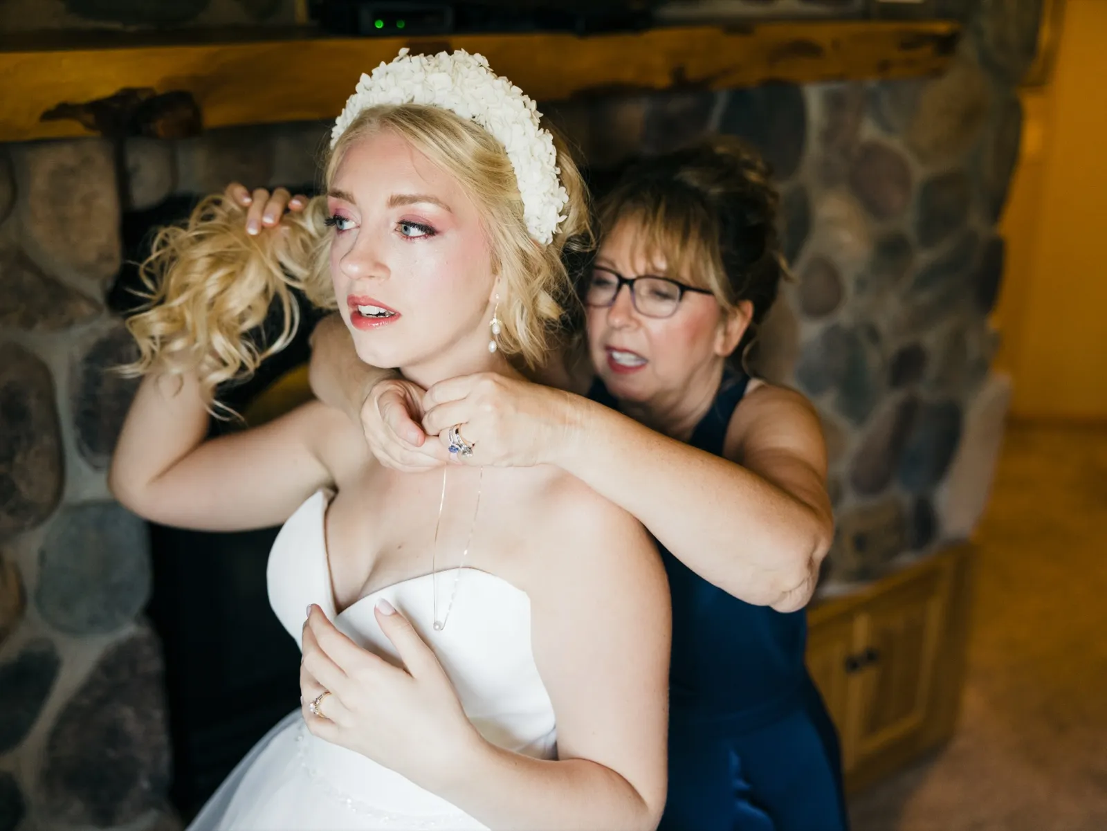 The bride lifts her hair as her mother clasps a delicate necklace at her neck in front of a stone fireplace. — Tim Larsen Photography, Brainerd Lakes MN