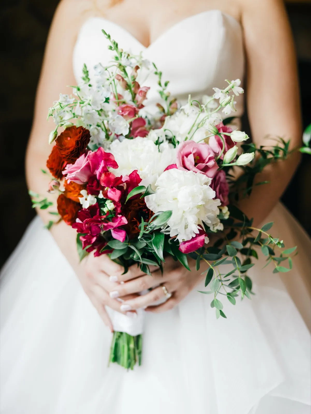 The bride holds a lush bouquet of white peonies, pink lisianthus, red ranunculus, and trailing eucalyptus against her strapless white gown. — Tim Larsen Photography, Brainerd Lakes MN