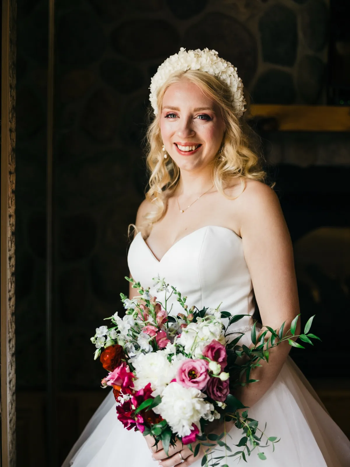 The bride smiles at the camera in a strapless gown and white floral crown, holding a jewel-toned bouquet of pink, red, and white blooms in front of a dark stone wall. — Tim Larsen Photography, Brainerd Lakes MN