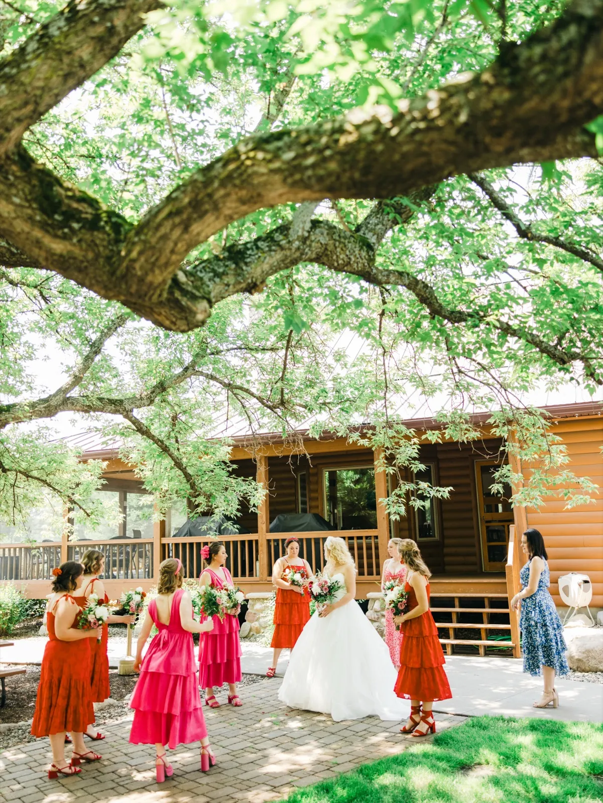 A bride in a white gown stands with her bridal party in pink and red dresses on a paver patio beneath a sprawling oak tree outside a log cabin lodge. — Tim Larsen Photography, Brainerd Lakes MN