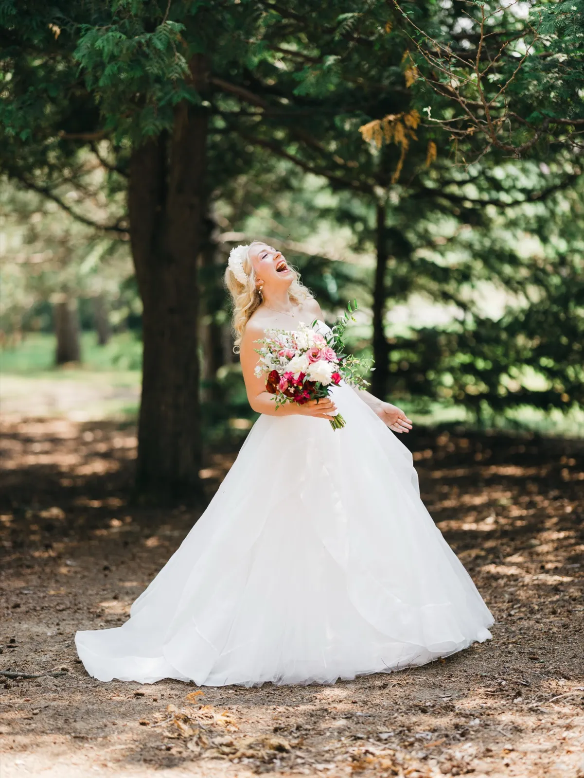 The bride throws her head back laughing while holding a pink and white bouquet, her full tulle gown fanning out around her in a sunlit pine grove. — Tim Larsen Photography, Brainerd Lakes MN