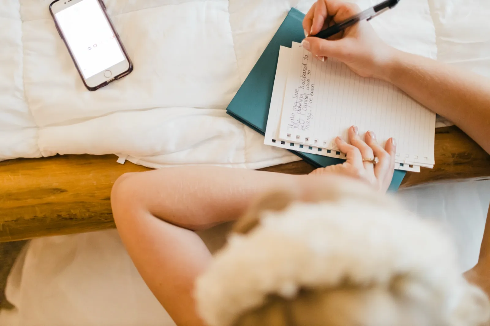 Overhead view of the bride seated on a bed, writing handwritten vows to her future husband on lined notecards beside her phone and bouquet. — Tim Larsen Photography, Brainerd Lakes MN