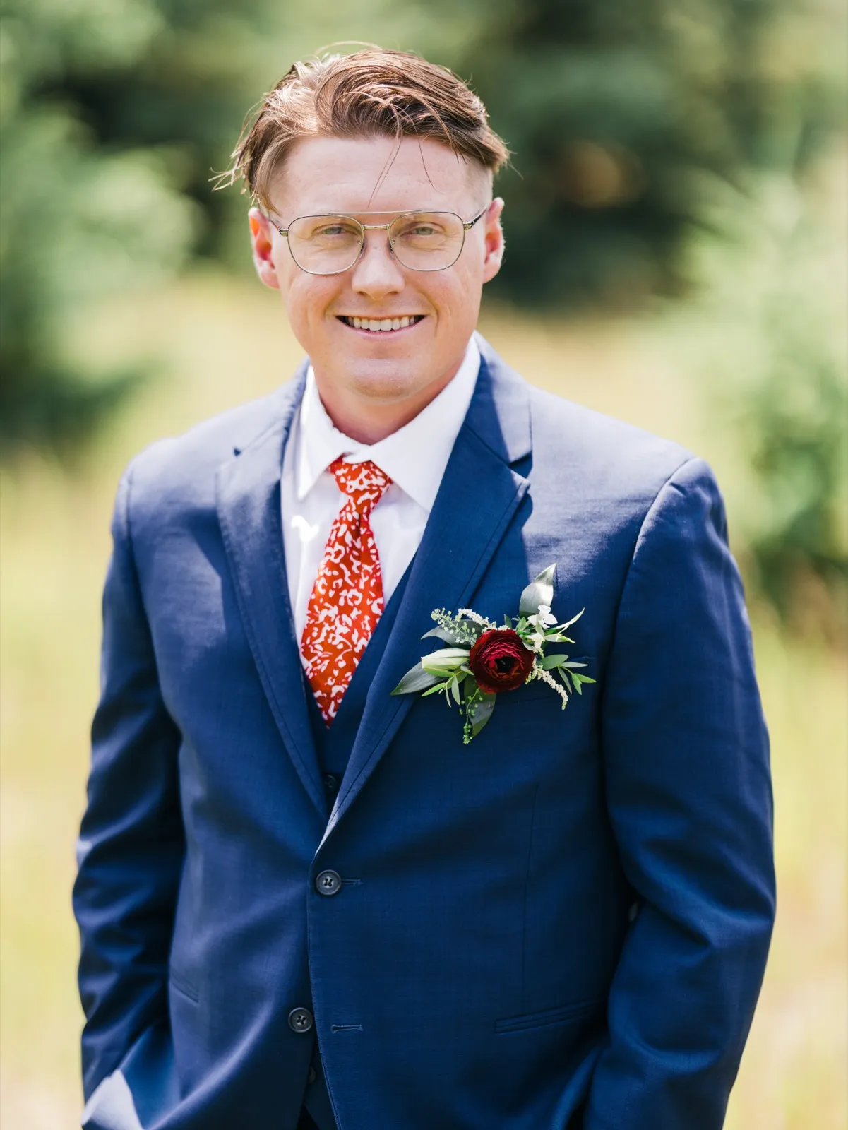 The groom stands in a field in a navy suit and red patterned tie, smiling warmly at the camera with a deep red ranunculus boutonniere on his lapel. — Tim Larsen Photography, Brainerd Lakes MN