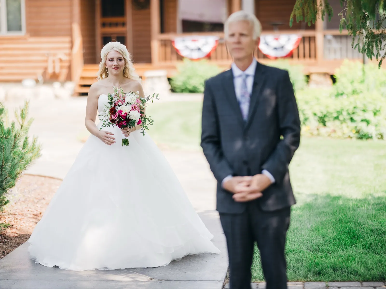 The bride approaches her groom from behind, holding a pink and burgundy bouquet, while he waits with his back turned on a walkway in front of a wood-sided lodge. — Tim Larsen Photography, Brainerd Lakes MN