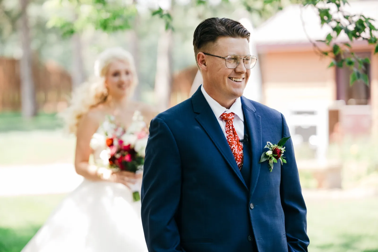The groom smiles broadly while the bride approaches behind him out of focus, holding a red and white bouquet on a sunlit garden path. — Tim Larsen Photography, Brainerd Lakes MN
