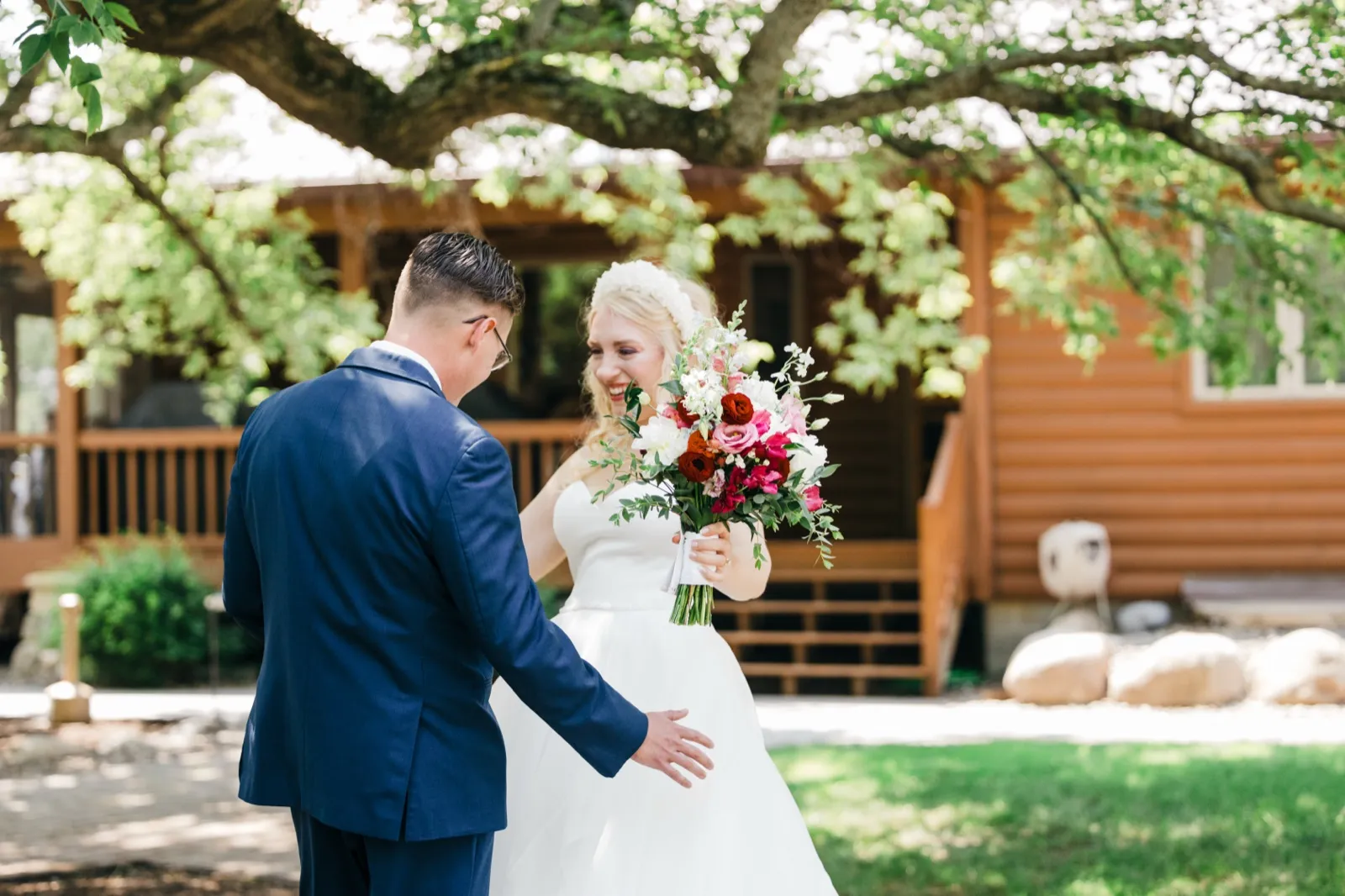 The bride laughs and beams while holding a bright bouquet as the groom reaches toward her during a first look outside the Pine Peaks Event Center lodge. — Tim Larsen Photography, Brainerd Lakes MN