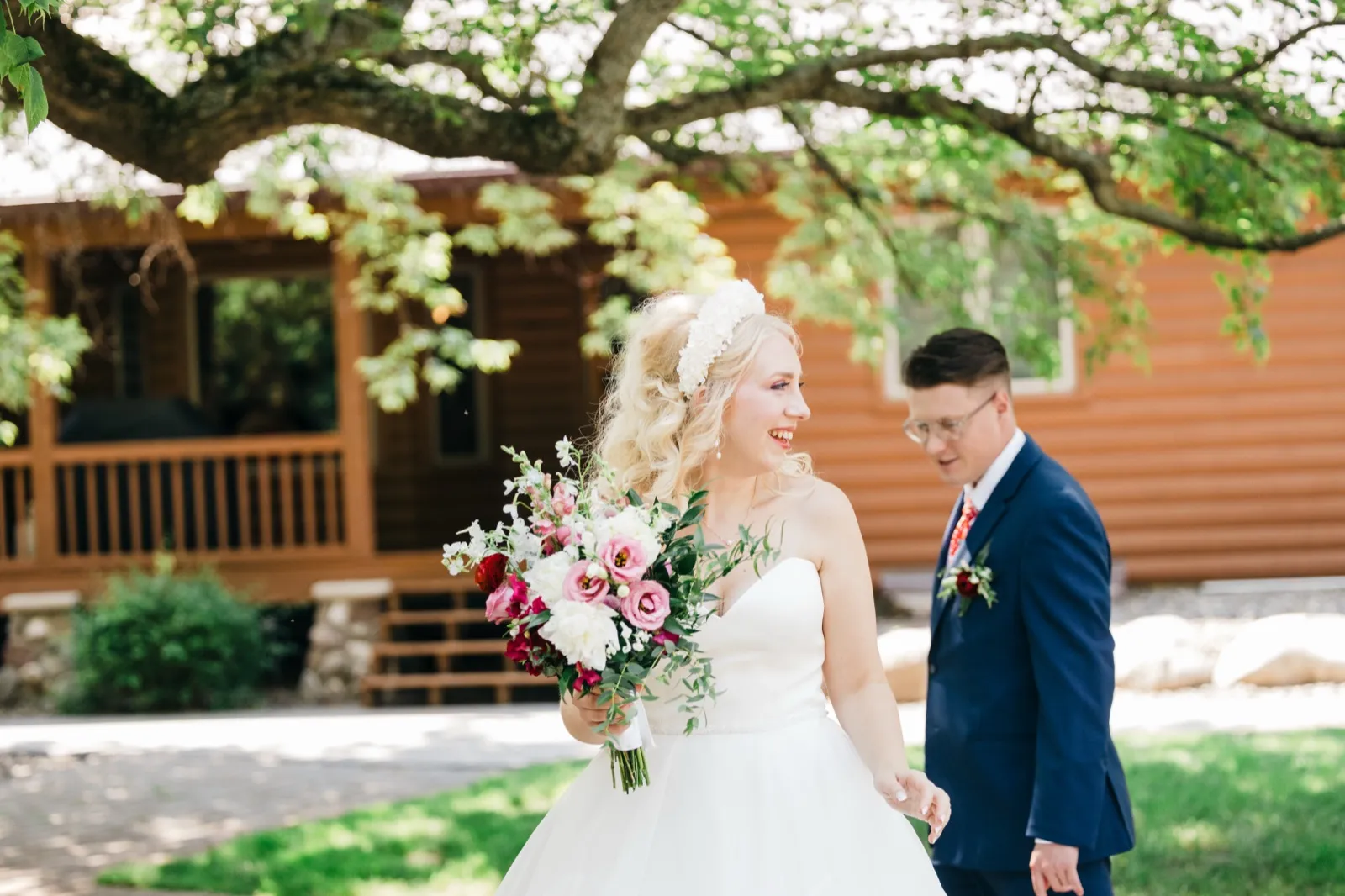 The bride laughs while holding a pink and white bouquet as the groom walks behind her in front of a wood-sided lodge framed by an oak canopy. — Tim Larsen Photography, Brainerd Lakes MN
