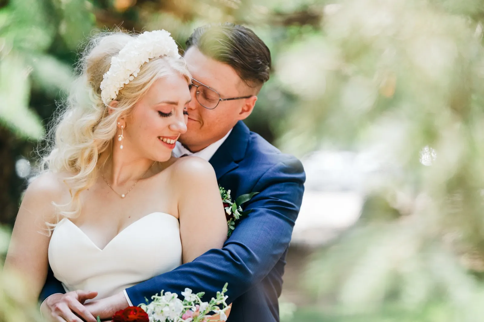 The groom nuzzles into the bride's cheek as she smiles with closed eyes, framed by soft green foliage in a sunlit forest clearing. — Tim Larsen Photography, Brainerd Lakes MN