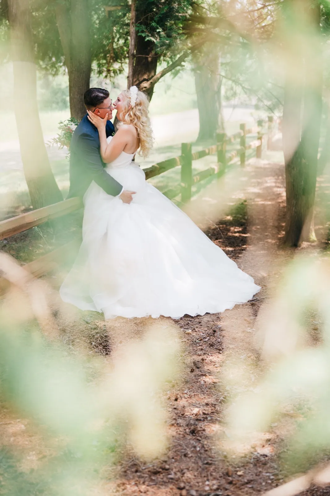 The bride and groom lean in close on a tree-lined path beside a split-rail fence at Pine Peaks Event Center, framed by soft out-of-focus foliage in the foreground. — Tim Larsen Photography, Brainerd Lakes MN