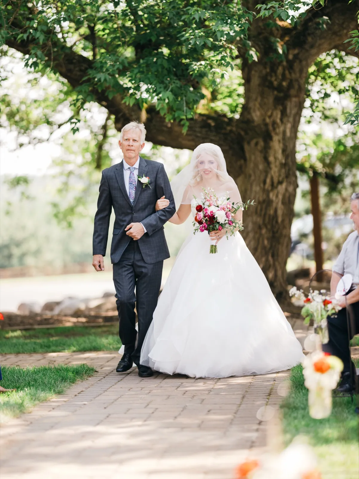 The bride walks down a brick ceremony aisle on her father's arm beneath a sprawling tree canopy, holding a bright pink and white bouquet. — Tim Larsen Photography, Brainerd Lakes MN
