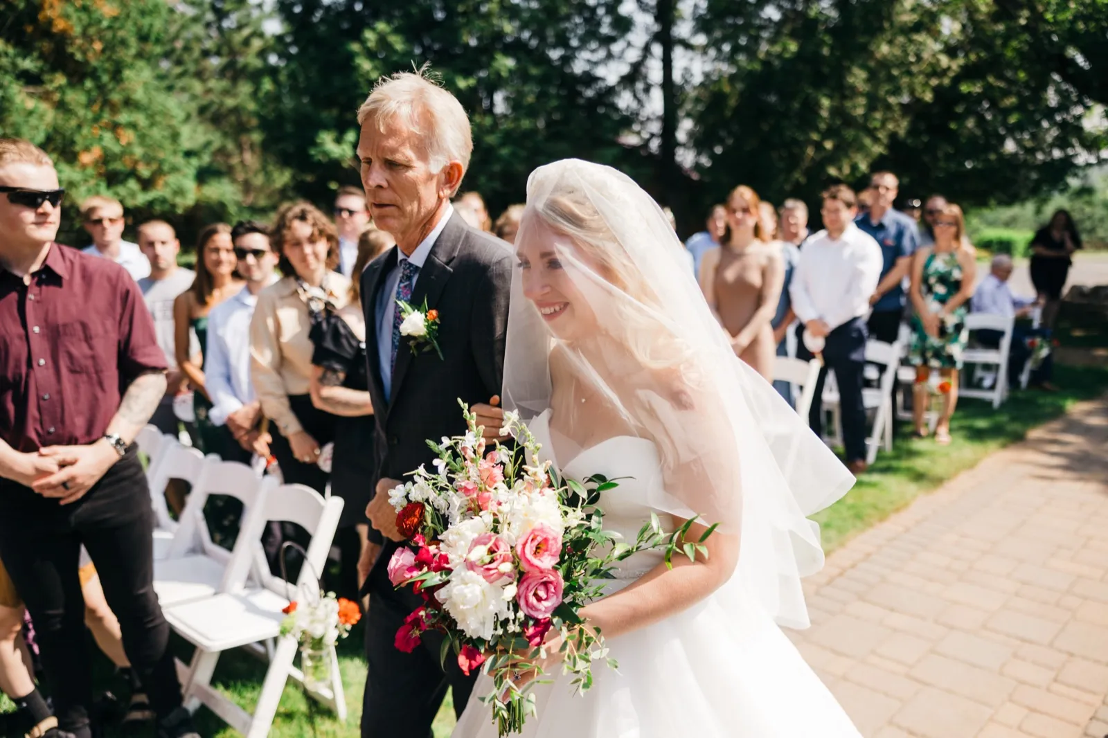 The bride smiles broadly under her veil while walking down the outdoor ceremony aisle on her father's arm, holding a large pink and red bouquet as guests stand on either side. — Tim Larsen Photography, Brainerd Lakes MN