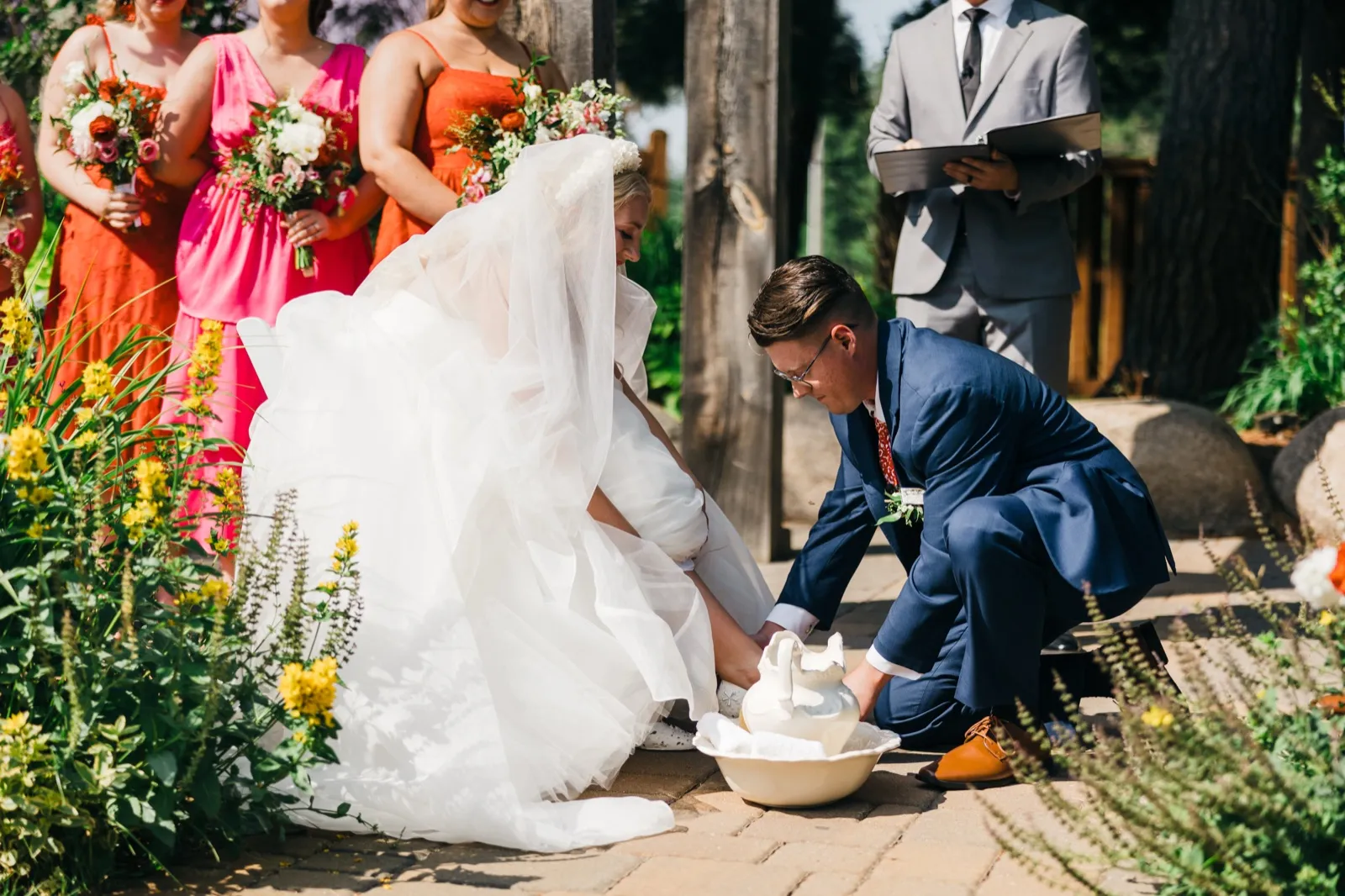 The groom kneels in a blue suit to wash the bride's feet from a ceramic basin during their outdoor ceremony, with bridesmaids in coral dresses and the officiant looking on. — Tim Larsen Photography, Brainerd Lakes MN