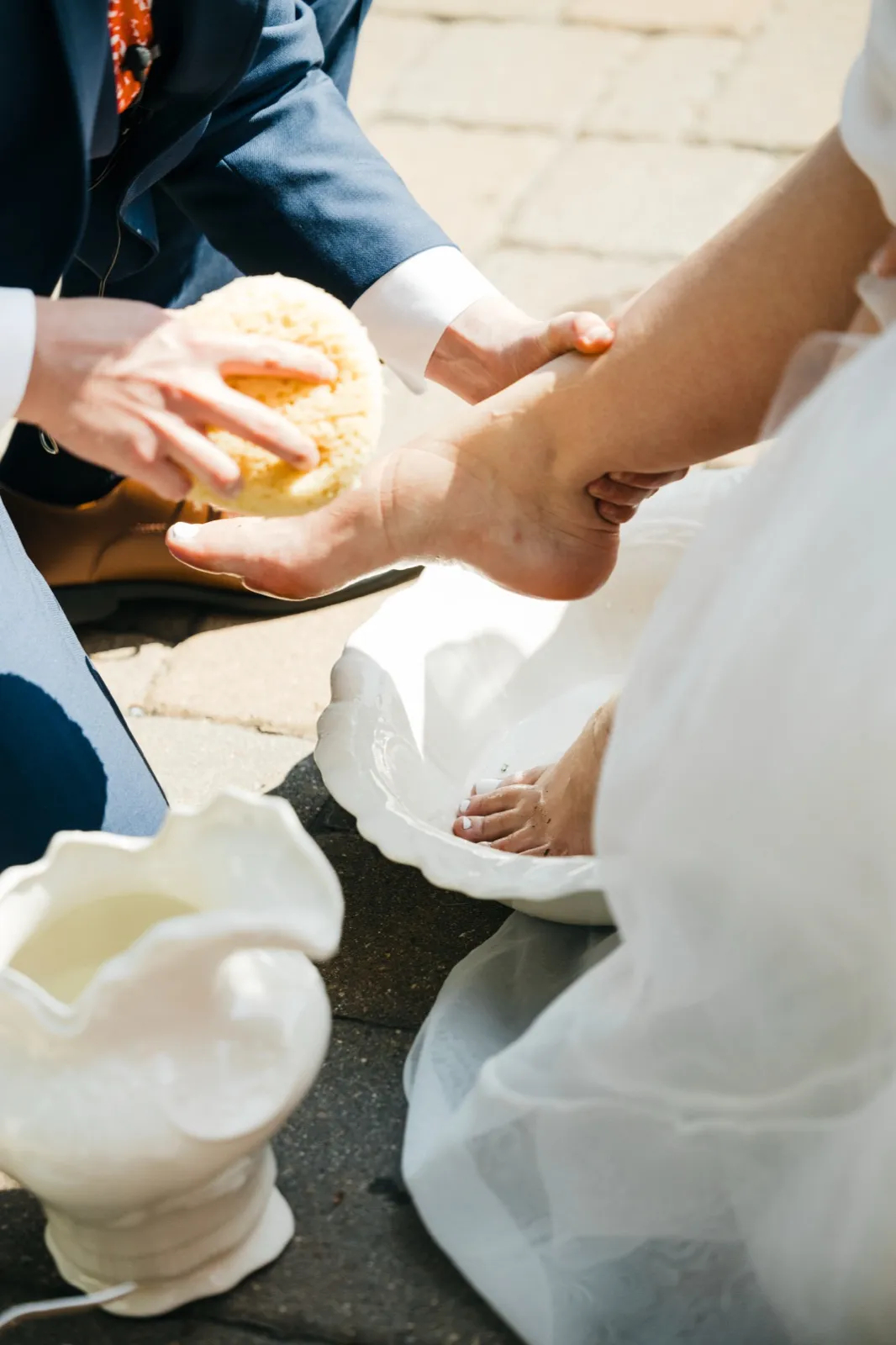 The groom kneels and uses a sponge to wash the bride's bare foot in a white ceramic basin during a foot-washing ritual at their ceremony.