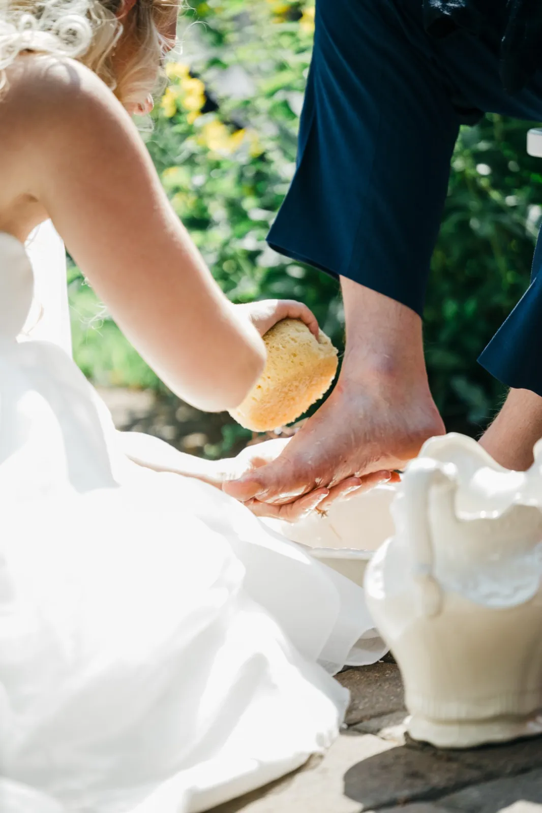 The bride washes the groom's bare foot with a yellow sponge over a white ceramic basin during an outdoor foot-washing ceremony.