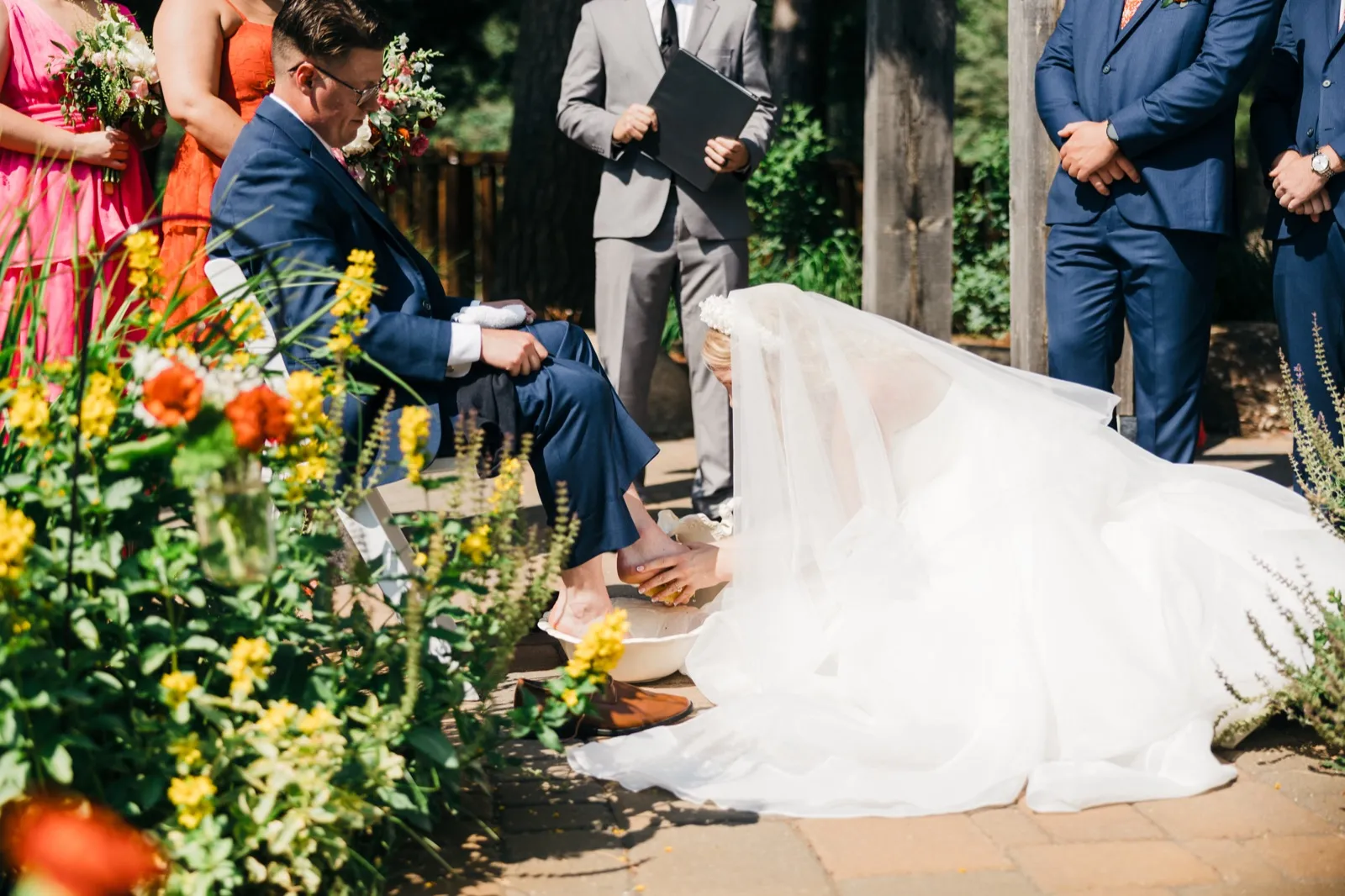 The bride kneels in her gown to wash the groom's feet during an outdoor ceremony, surrounded by the bridal party and framed by wildflowers in the foreground. — Tim Larsen Photography, Brainerd Lakes MN