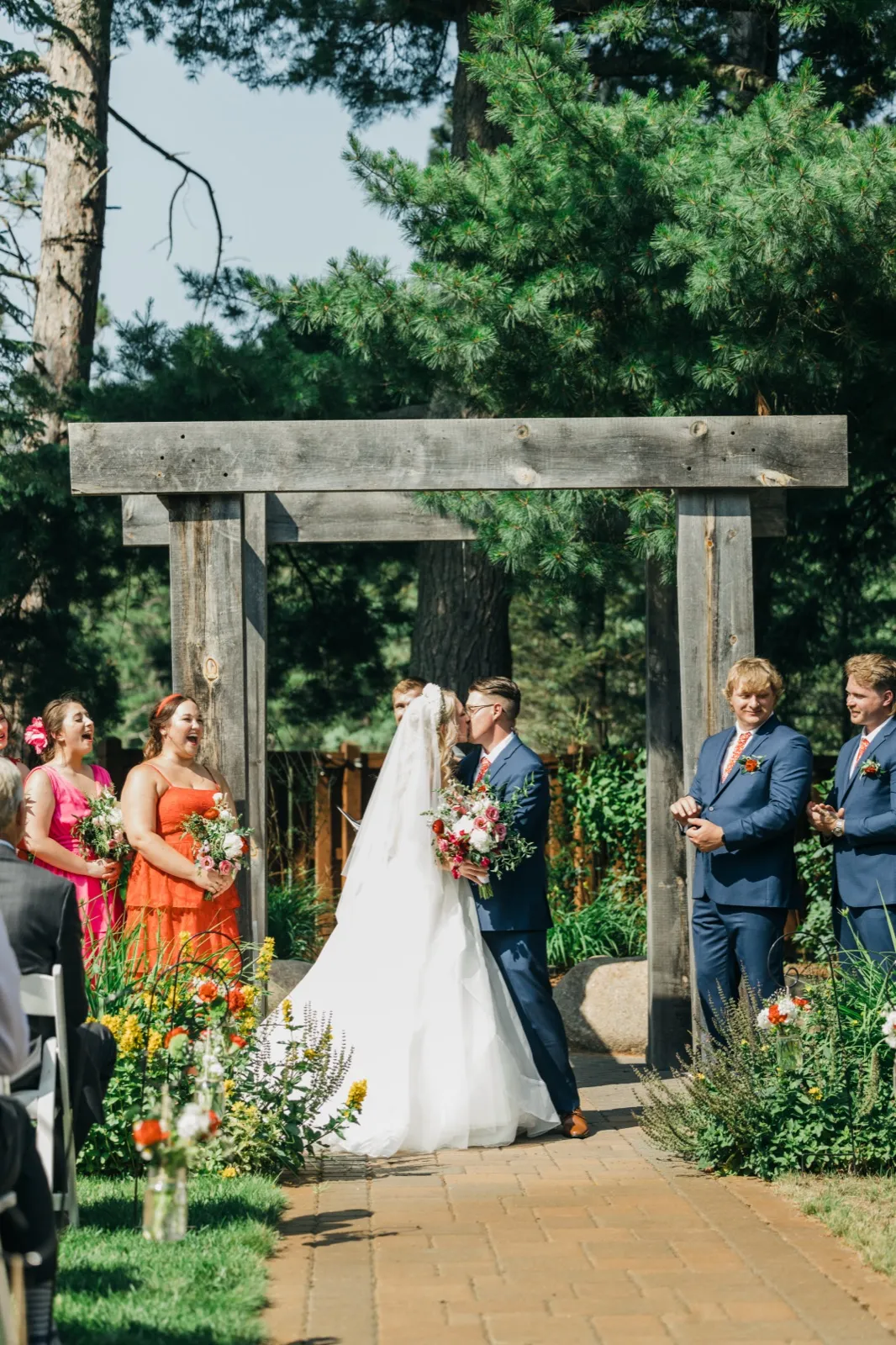 The bride and groom share their first kiss beneath a wooden arch in a pine forest while bridesmaids in pink and orange laugh and groomsmen in navy suits look on. — Tim Larsen Photography, Brainerd Lakes MN