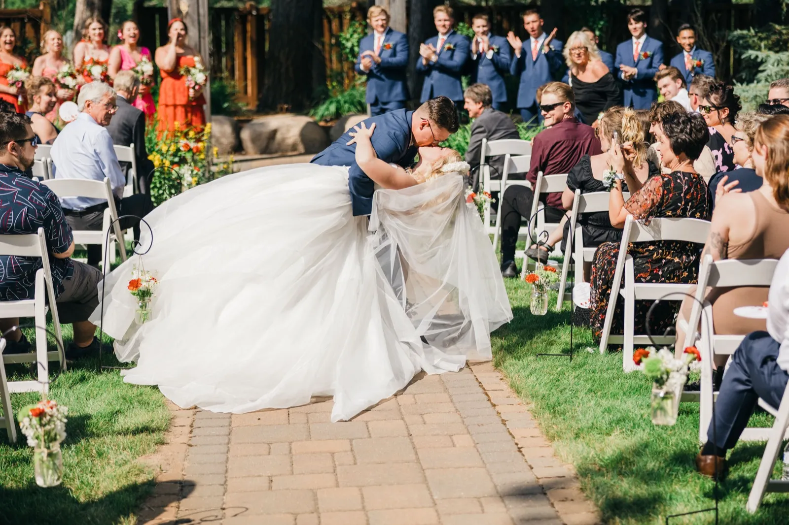 The groom dips the bride for a kiss in the middle of the aisle while the bridal party and seated guests laugh and applaud at an outdoor ceremony. — Tim Larsen Photography, Brainerd Lakes MN