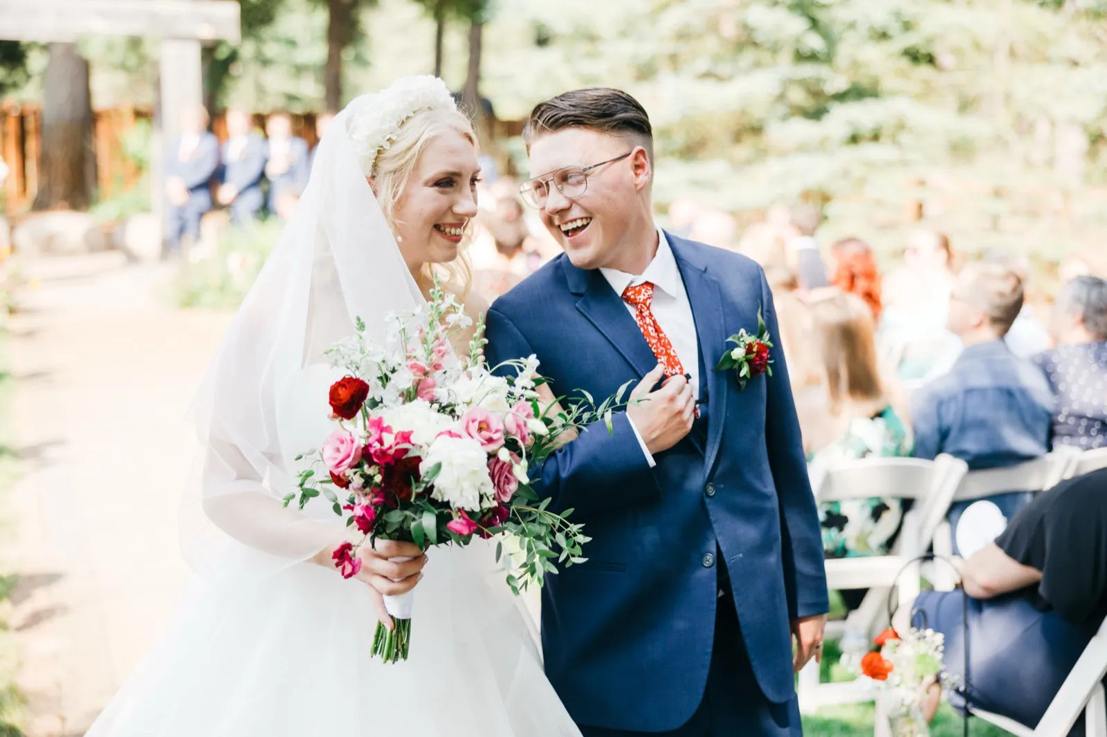 The bride and groom share a laughing glance as they walk down the forest aisle just after their ceremony, her bouquet of red and pink blooms held at her side. — Tim Larsen Photography, Brainerd Lakes MN