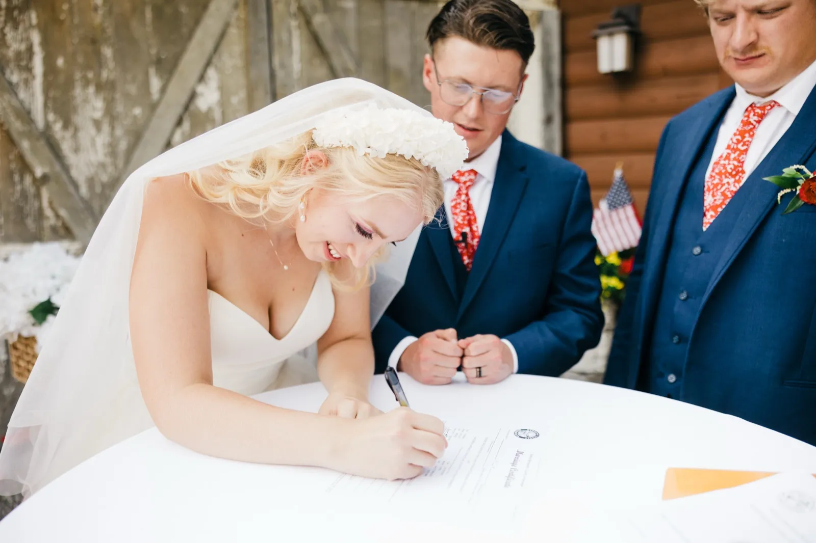 The bride, wearing a floral crown and long veil, smiles softly as she signs the marriage certificate while the groom watches from across the table. — Tim Larsen Photography, Brainerd Lakes MN