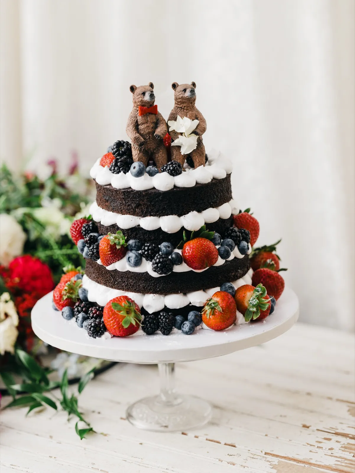 A three-tier naked chocolate wedding cake topped with two bear figurines and decorated with strawberries, blueberries, and blackberries on a white pedestal stand. — Tim Larsen Photography, Brainerd Lakes MN