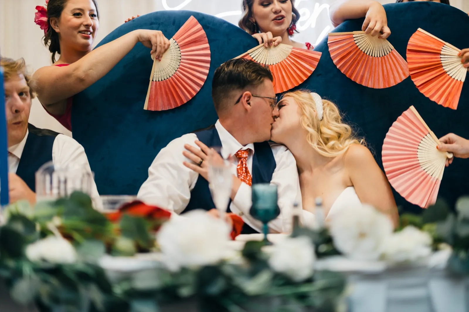 The bride and groom share a kiss at the head table while bridesmaids on either side hold up coral paper fans behind them. — Tim Larsen Photography, Brainerd Lakes MN
